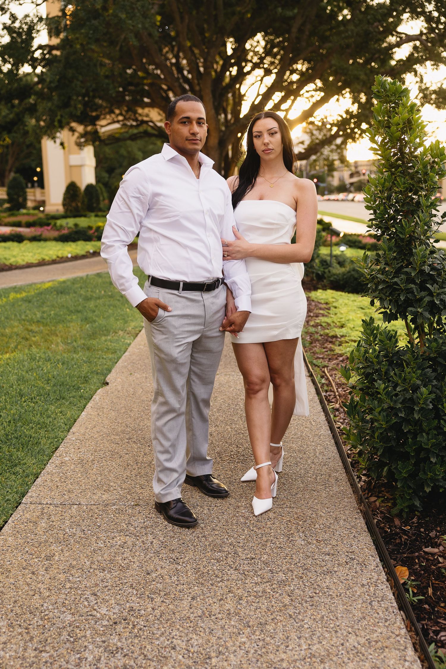 Stylish couple posing on garden walkway at sunset, man in white shirt with woman in white strapless mini dress.