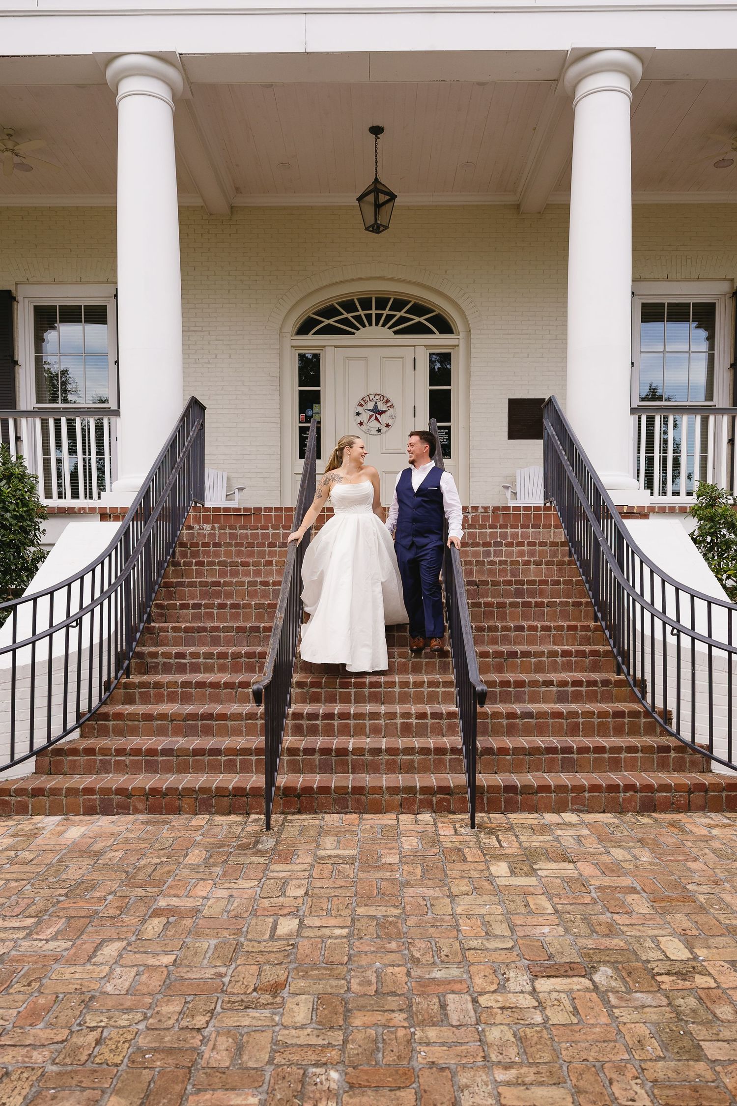 Elegant bride and groom walking down brick stairs of a white colonial wedding venue with tall columns.