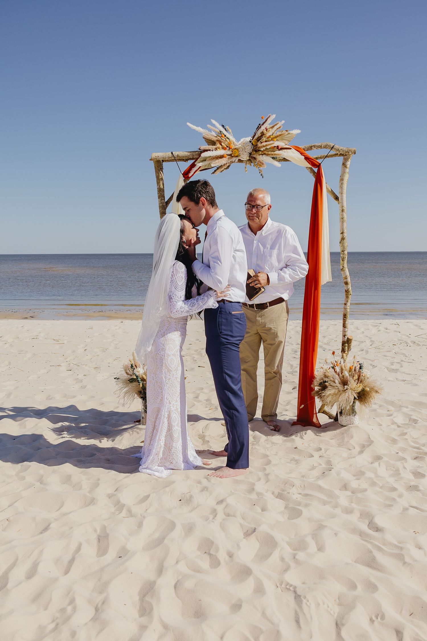 Bride and groom kissing under rustic wooden arch with orange fabric and dried florals at beach wedding ceremony.