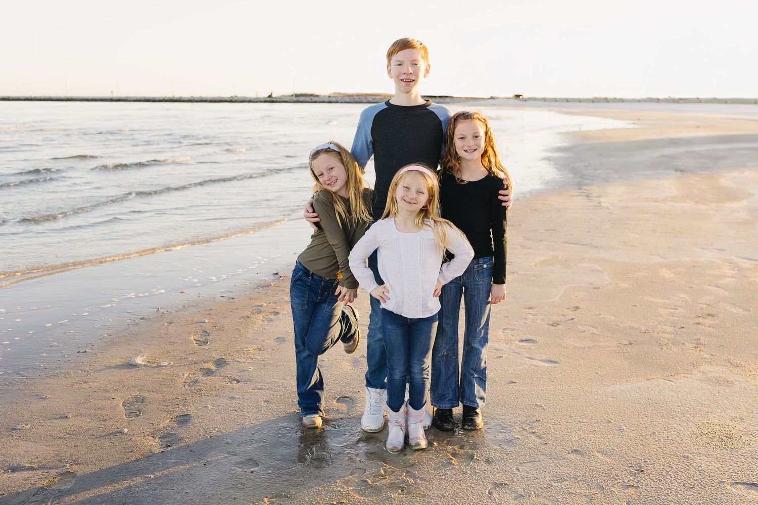 Four siblings pose together smiling on a sandy beach with calm ocean water behind them.