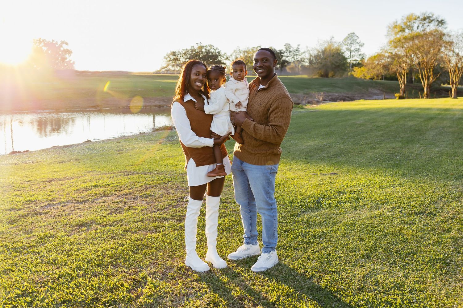A smiling family of three posing together on green grass with a glowing sunset behind them.