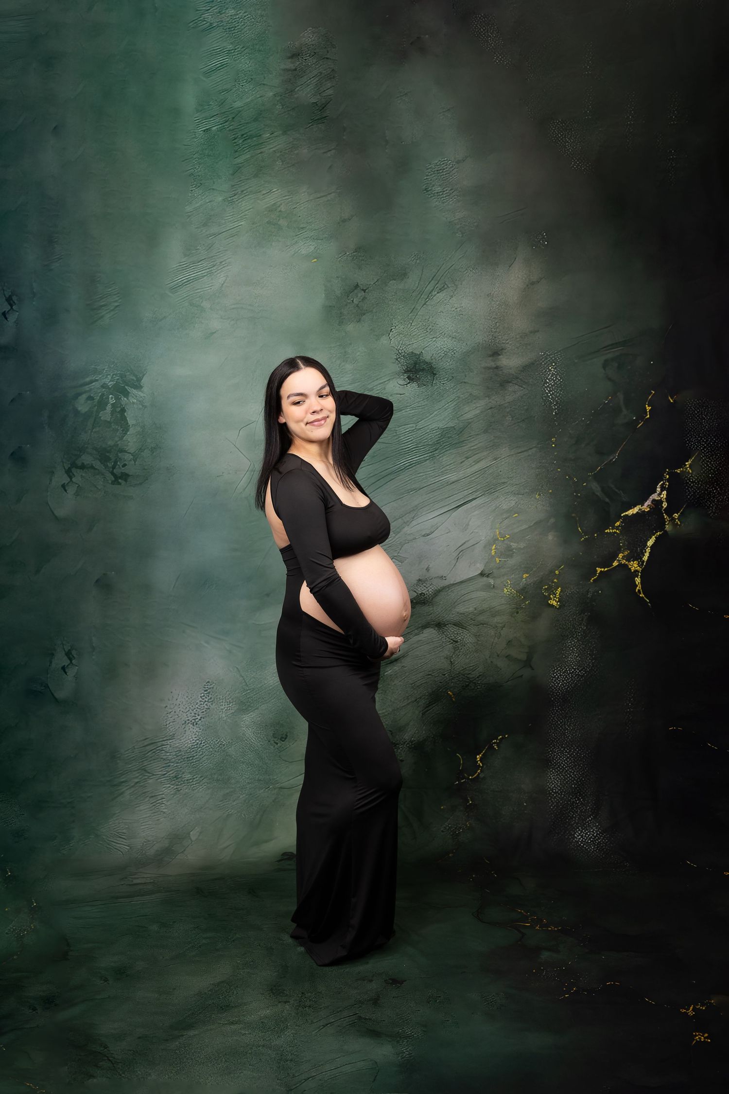 Pregnant woman in black form-fitting dress posing against a dark green textured studio backdrop.