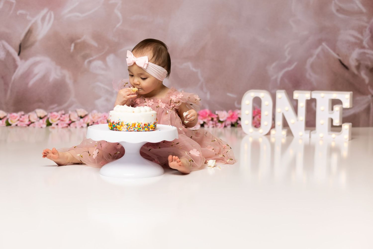 Baby girl in pink dress leaning over first birthday cake during a cake smash photo session.
