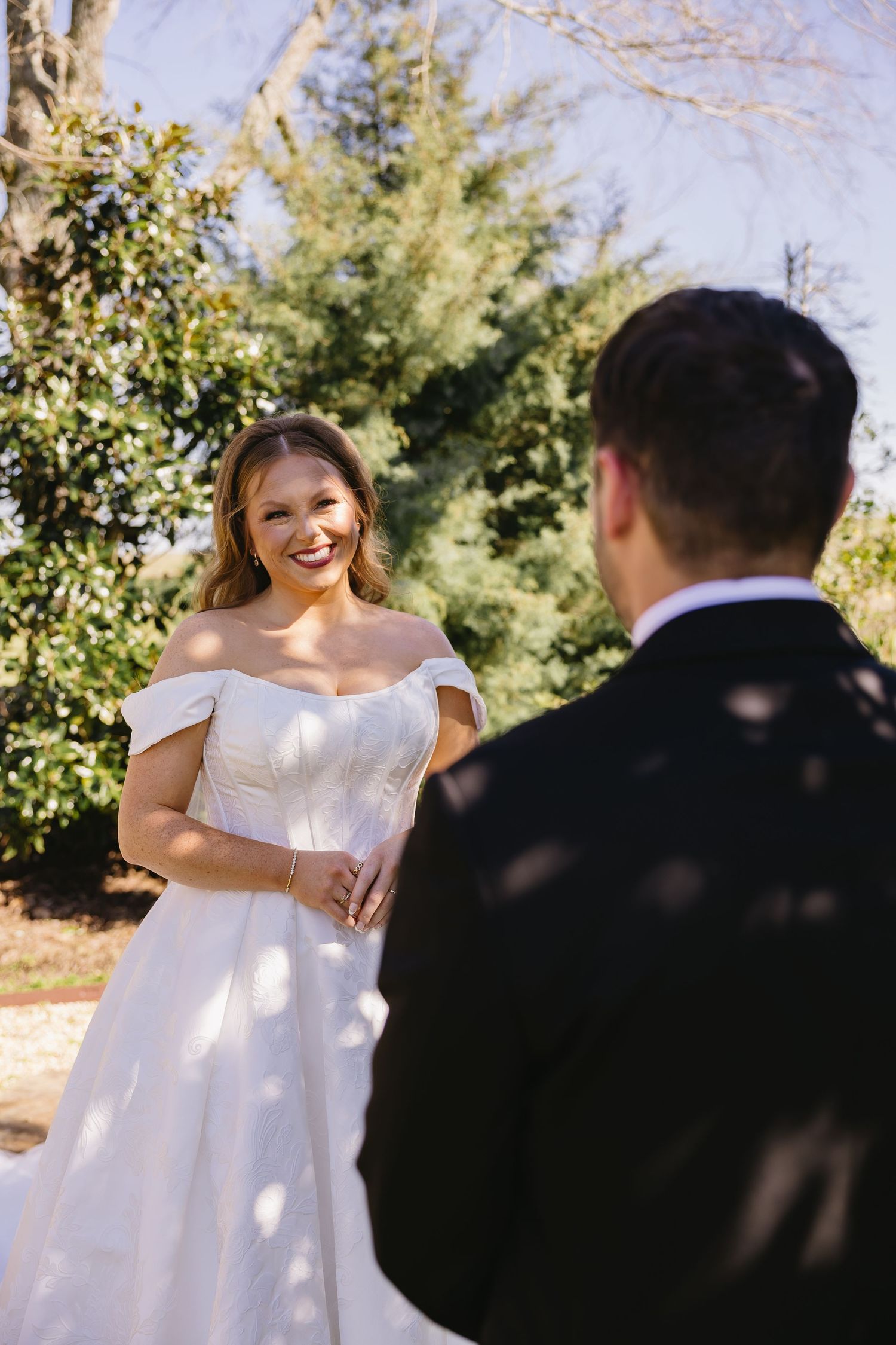 Smiling bride in an off-shoulder white gown during first look with groom in black tuxedo outdoors.