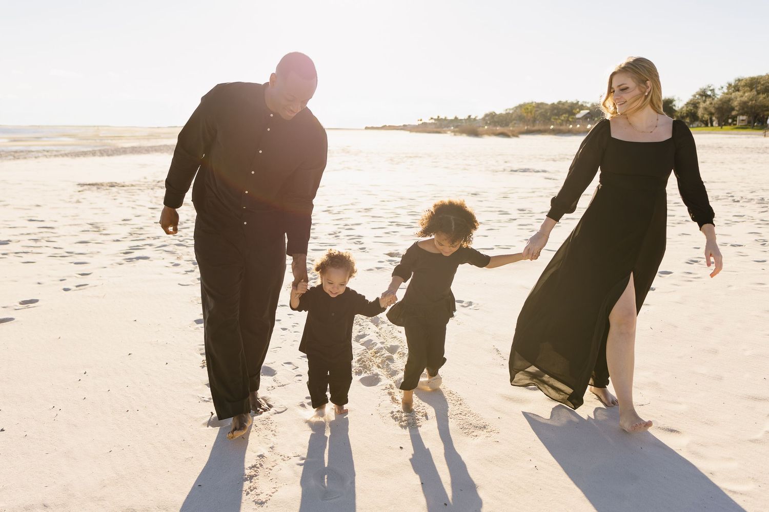 Joyful family in black outfits holding hands while walking on a sunny beach.