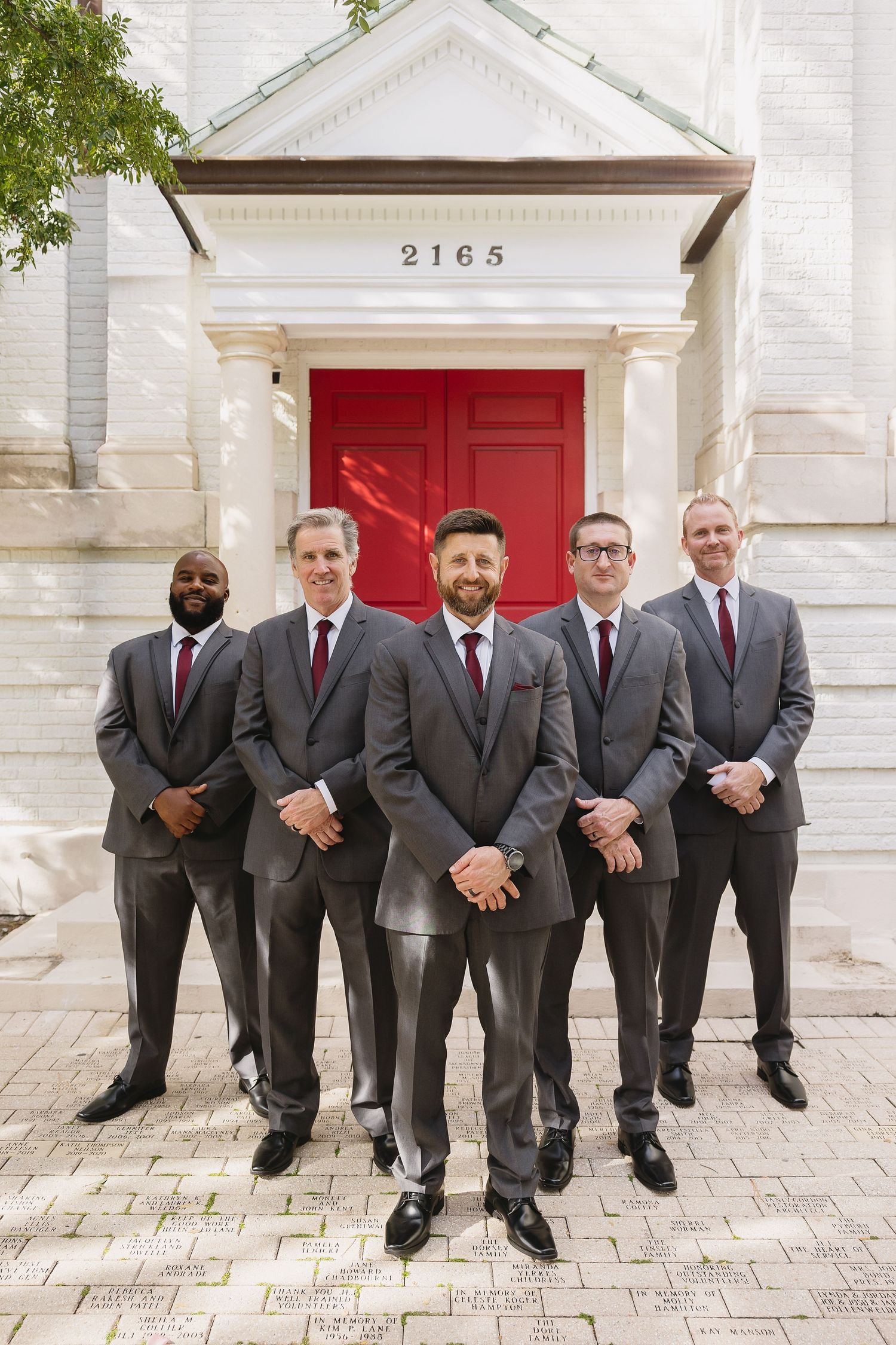 Groom and four groomsmen in gray suits posing before red church doors at 2165.
