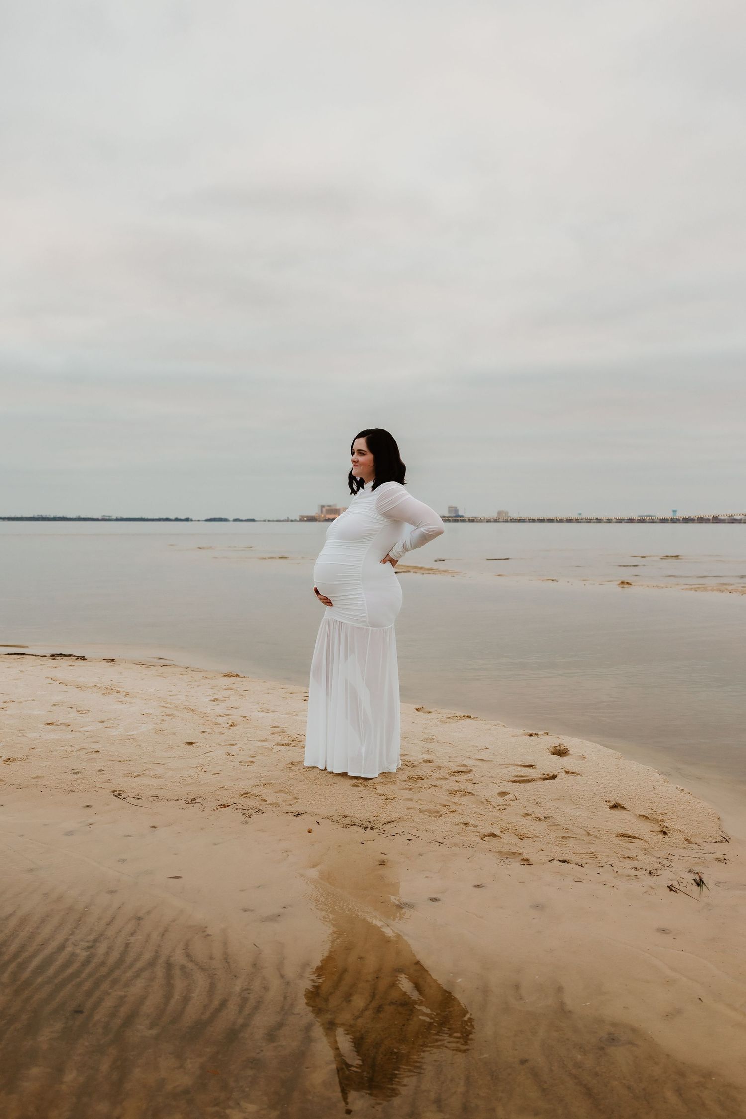 Pregnant woman in white sheer gown posing on sandy shoreline with calm water behind her.