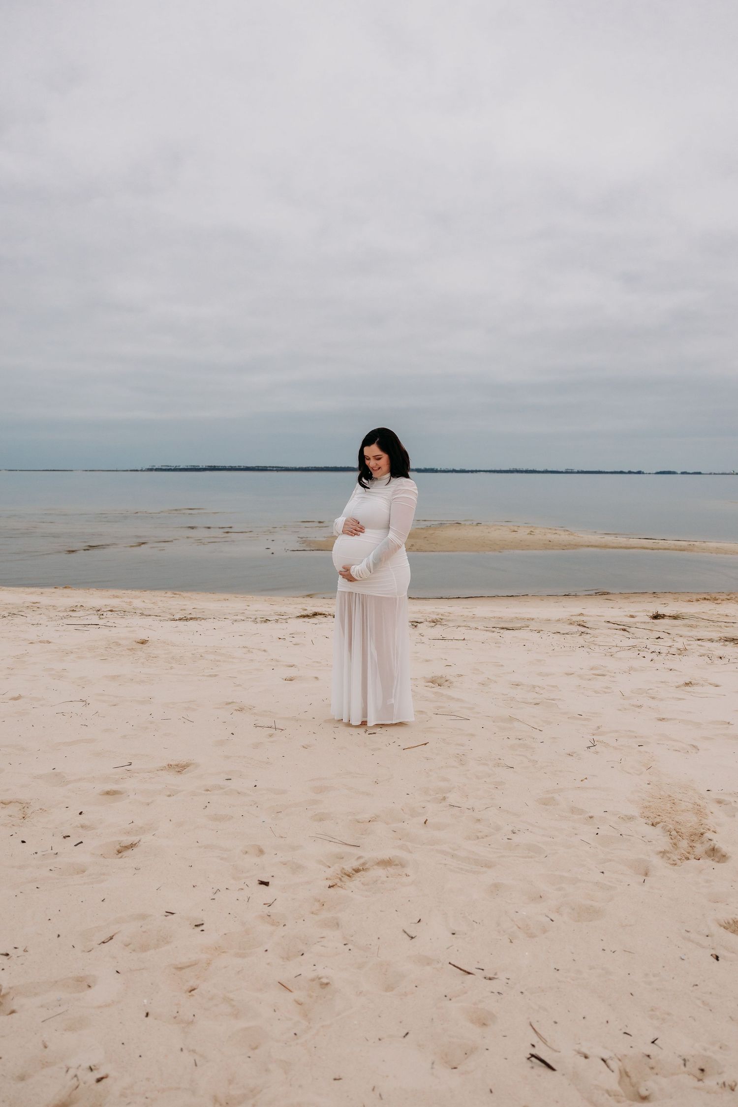 Pregnant woman in white maternity dress cradling belly on sandy beach.