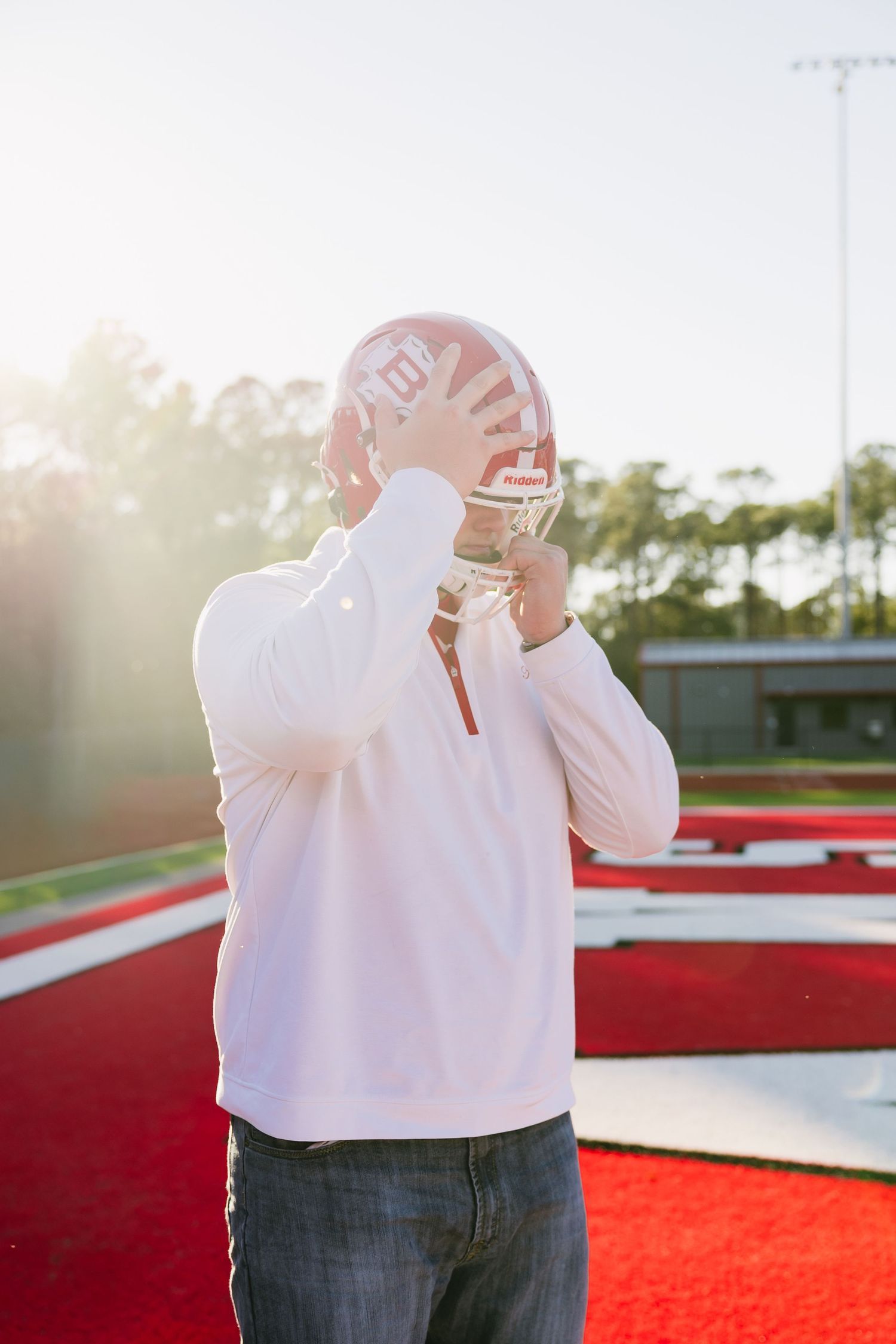 Football player in white jersey putting on red Riddell helmet on sunny field.