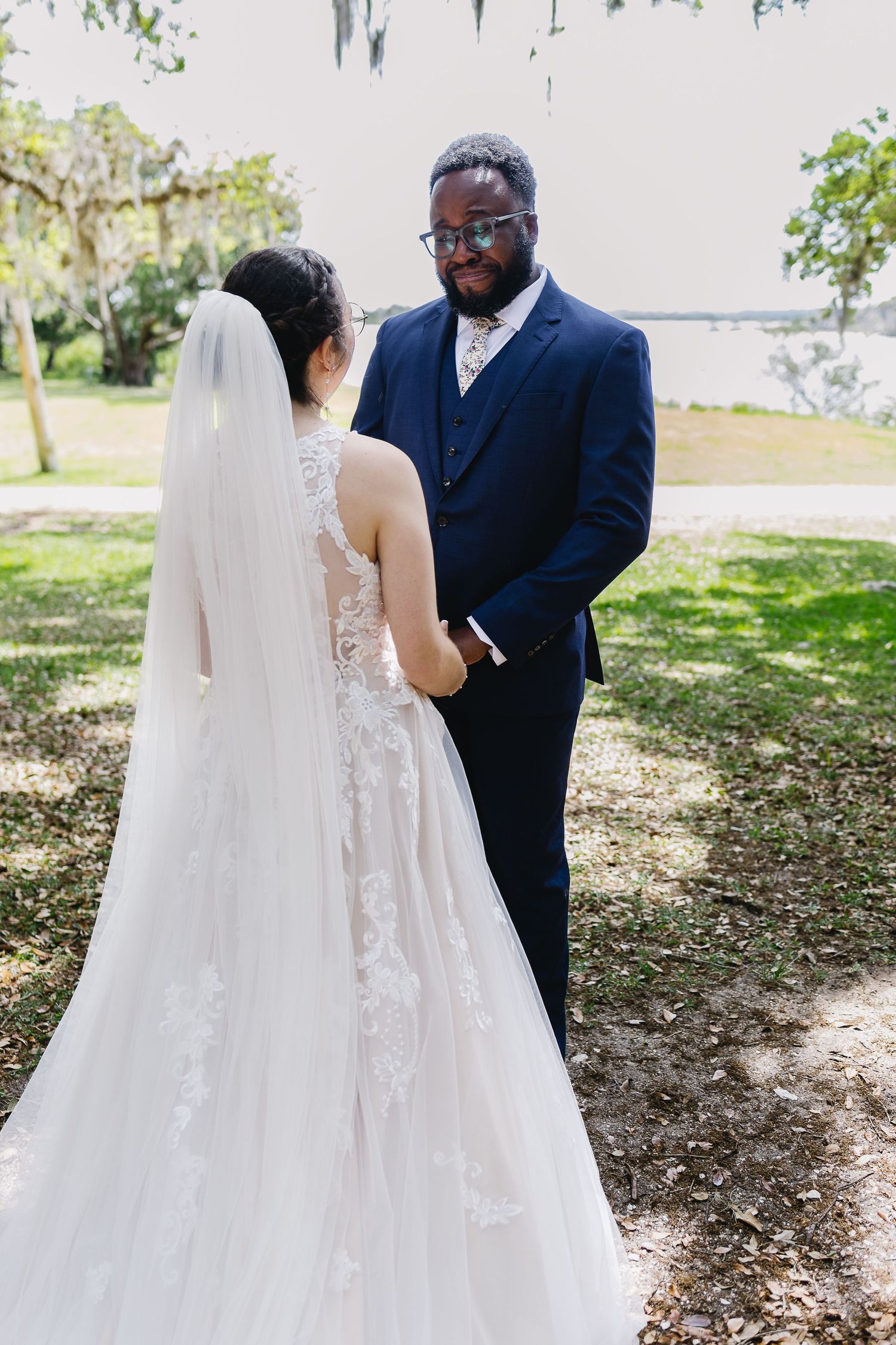 Bride in lace gown and groom in navy suit holding hands during outdoor wedding ceremony.
