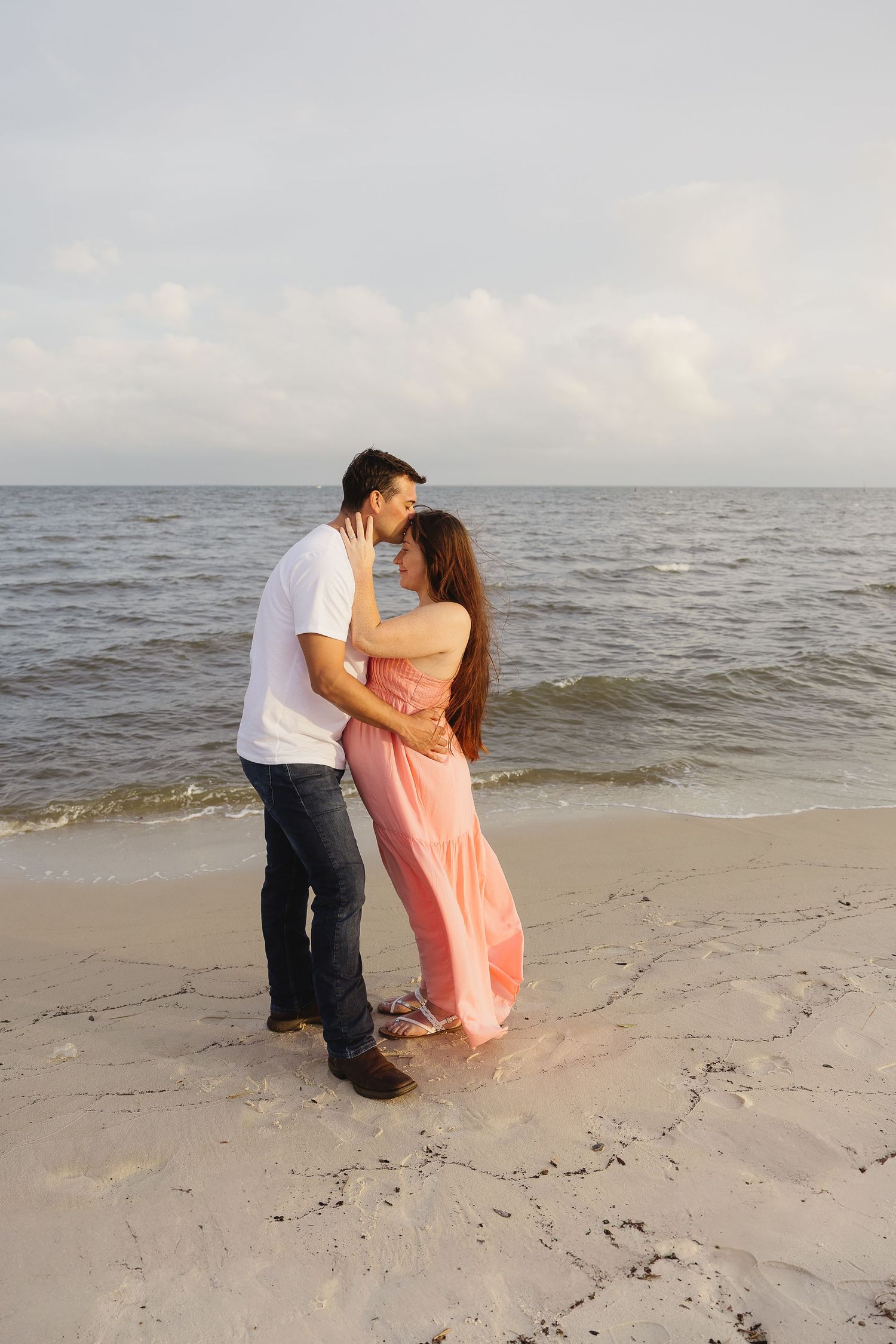 Expecting couple embracing on sandy beach shoreline, woman in flowing pink maternity dress.