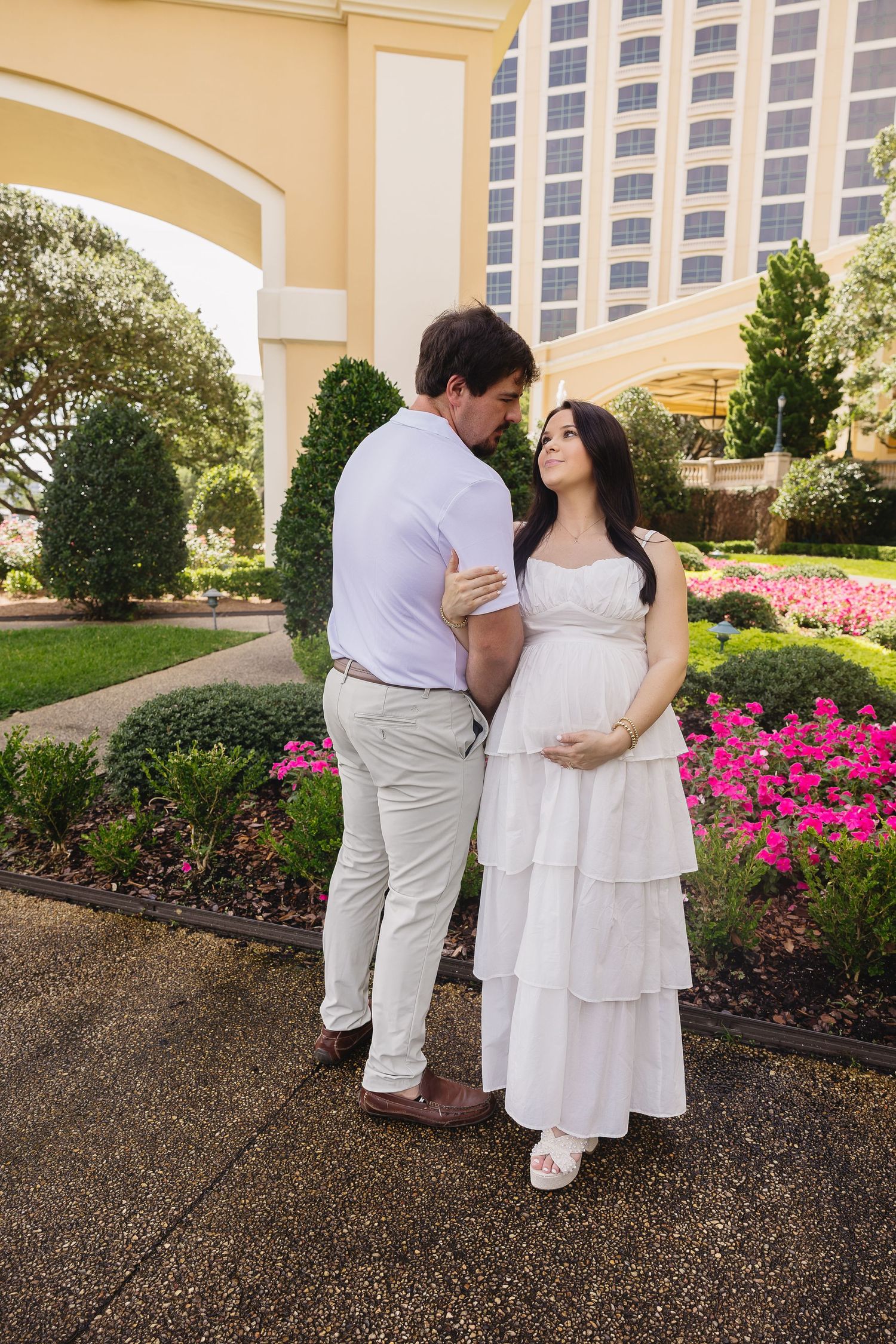 Couple in white outfits embracing in garden near building with pink flowers during maternity shoot.