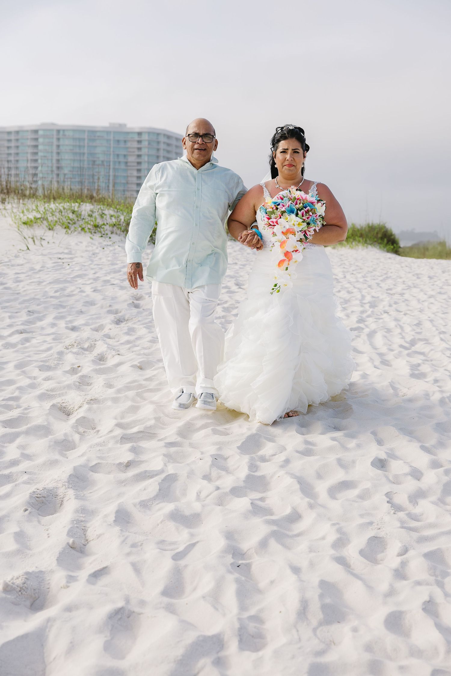 Bride in ruffled white gown holding colorful bouquet walks with groom on white sand beach.