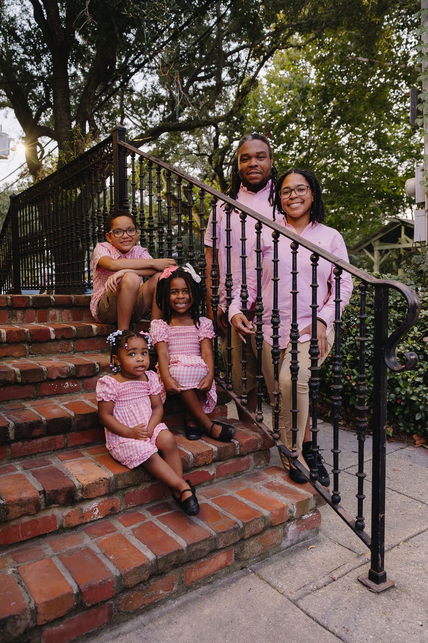 Family of five in pink outfits posing on brick stairs surrounded by lush green trees outdoors.