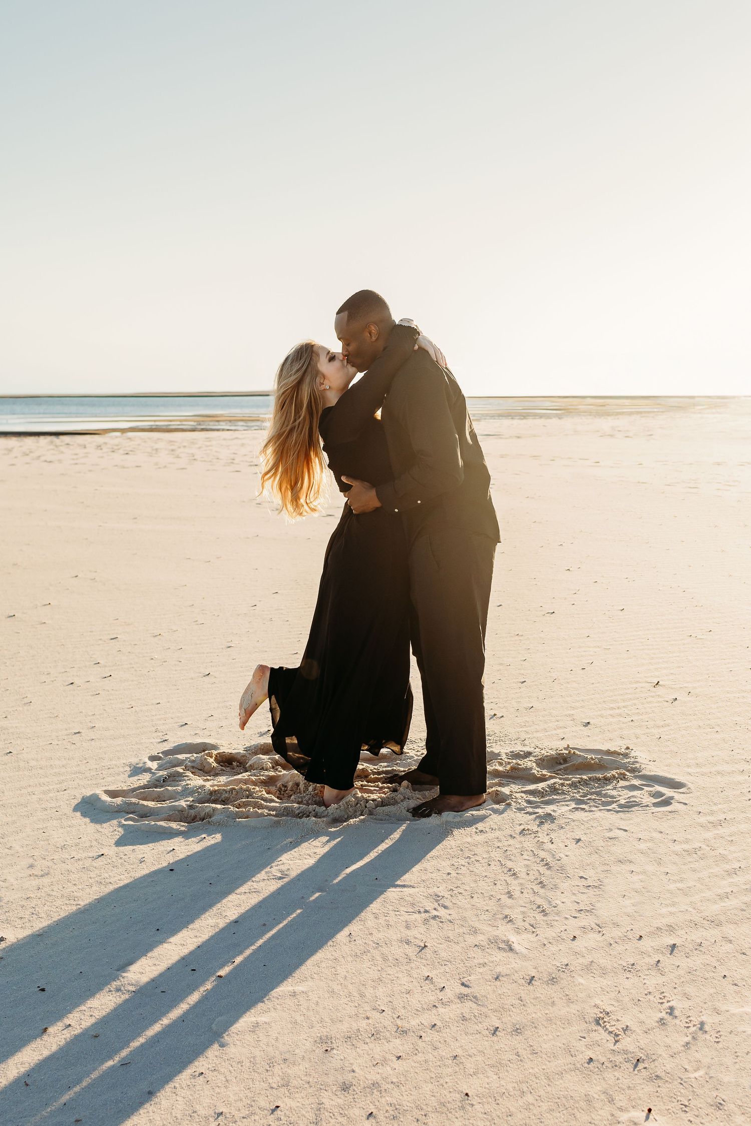 Interracial couple sharing a passionate kiss on a wide open sunlit sandy beach.