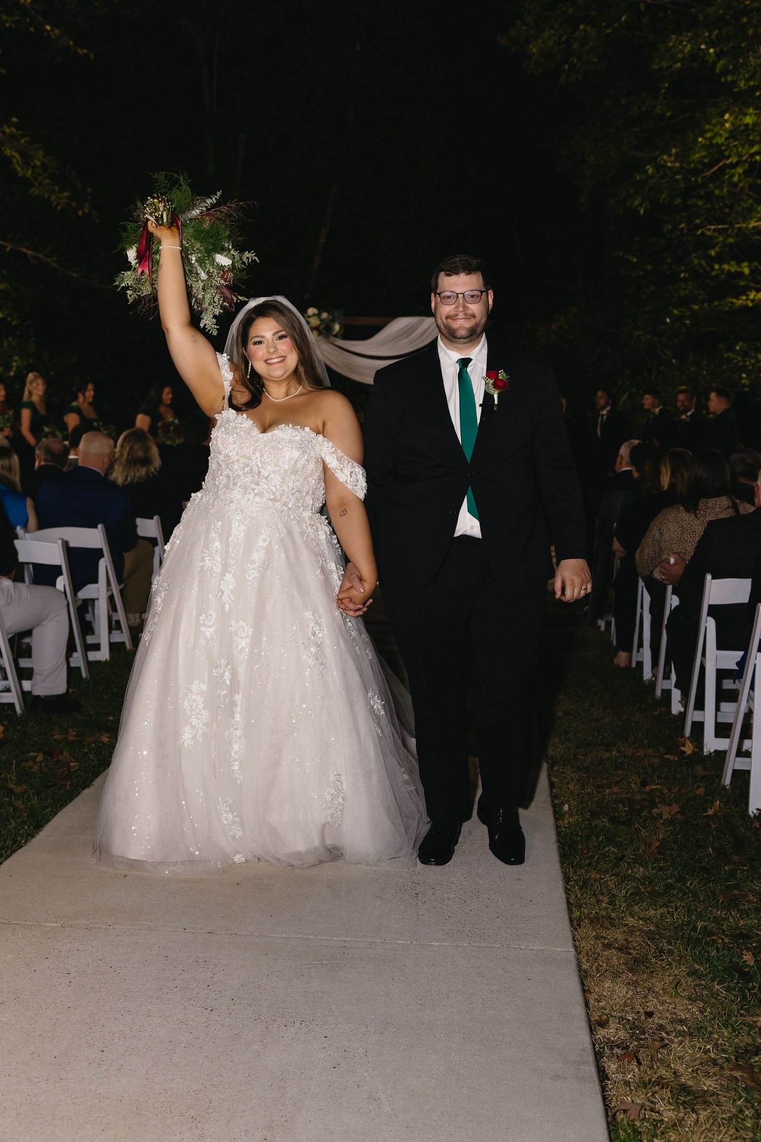 Bride raises bouquet triumphantly while walking down aisle with groom.