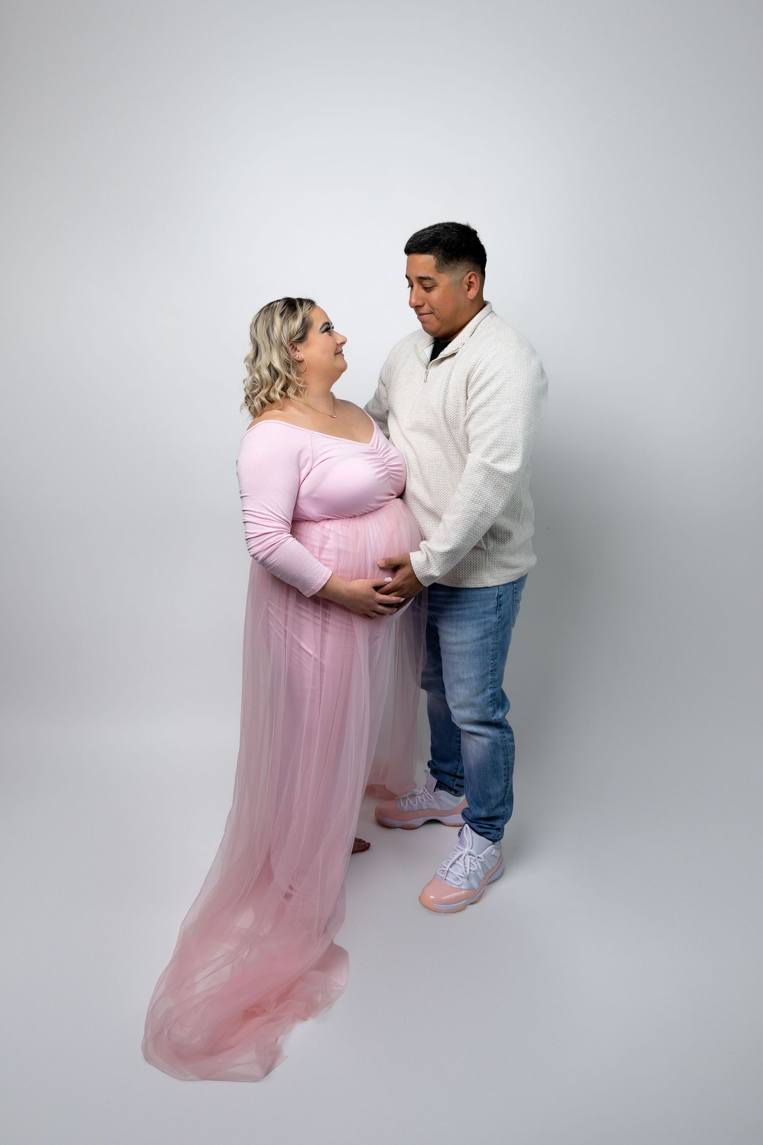 Pregnant woman in pink tulle maternity gown poses with partner in white sweater and jeans on white background.