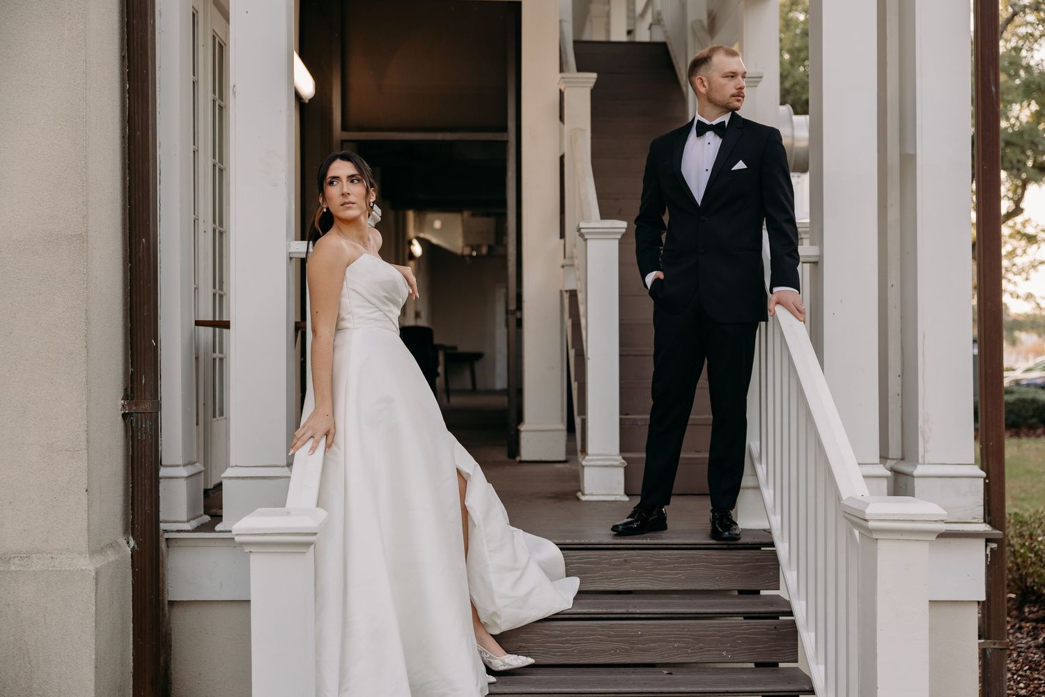Bride in a white ball gown and groom in black tuxedo pose on a classic columned porch staircase.
