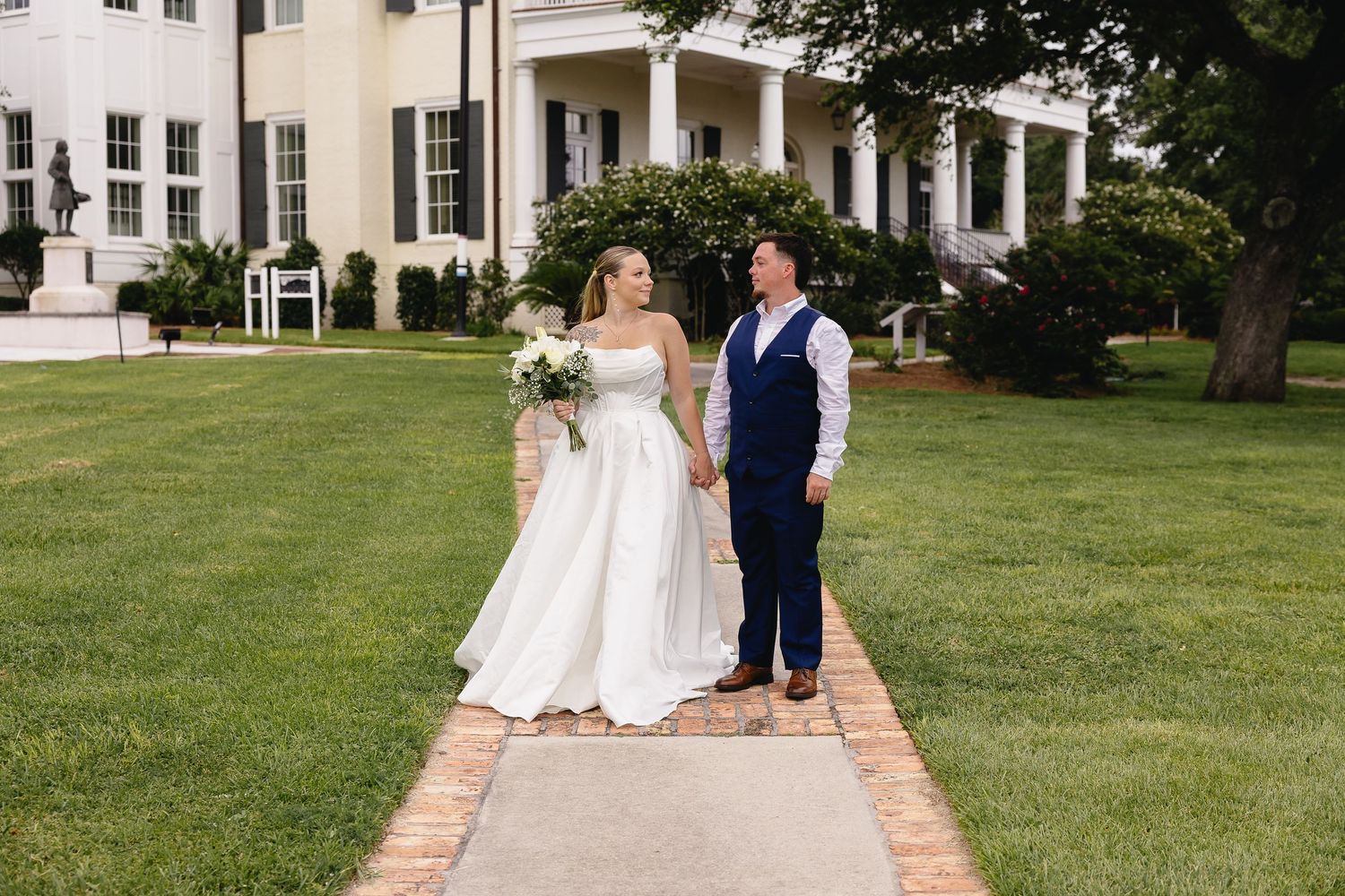 Bride and groom holding hands on a brick pathway in front of a grand white colonial mansion.