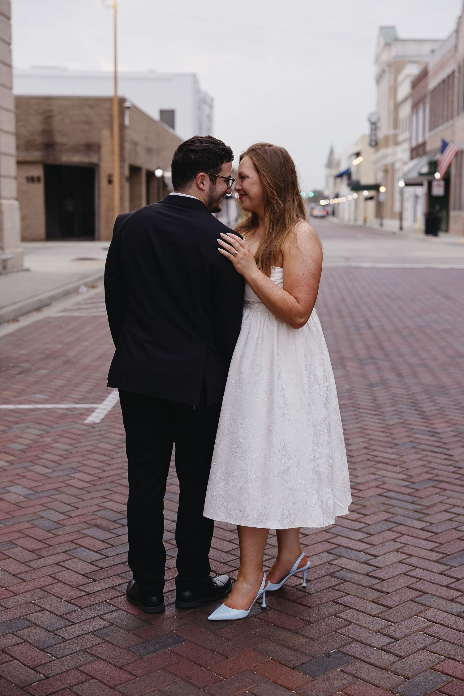 Bride and groom sharing an intimate moment on a brick street, bride in white midi dress and light blue heels.