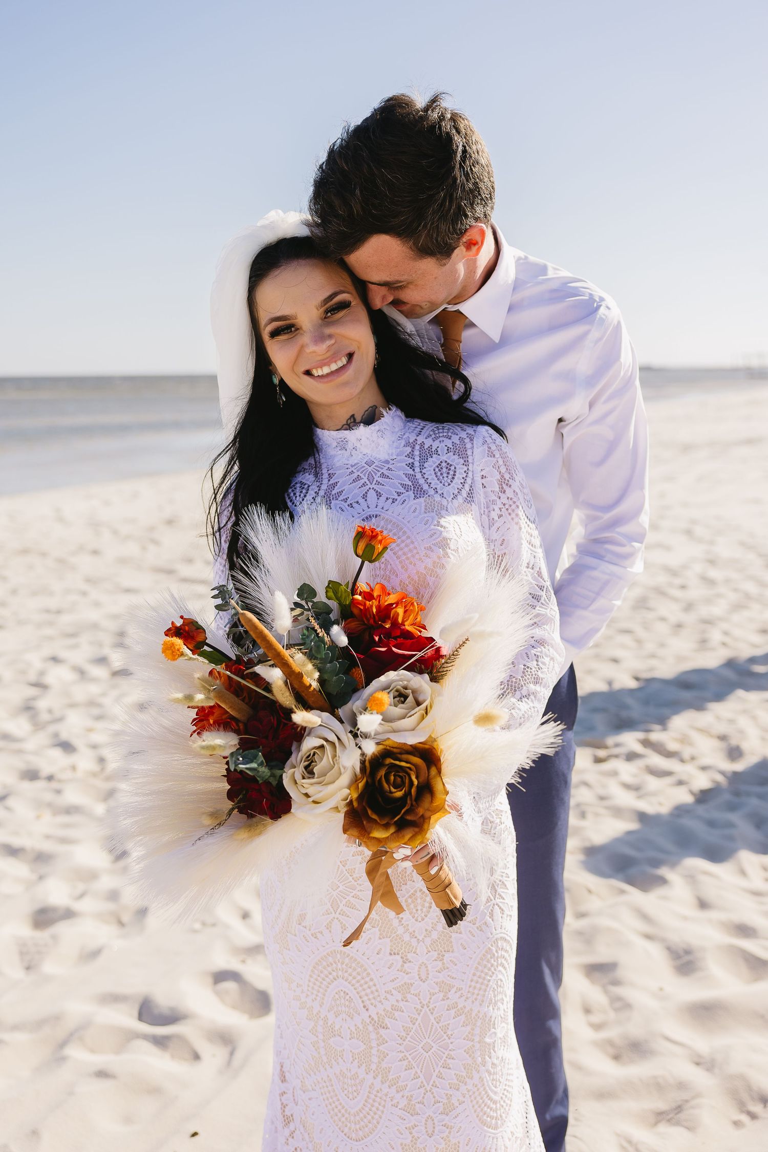 Smiling bride in lace gown holds boho bouquet as groom embraces her on sandy beach.