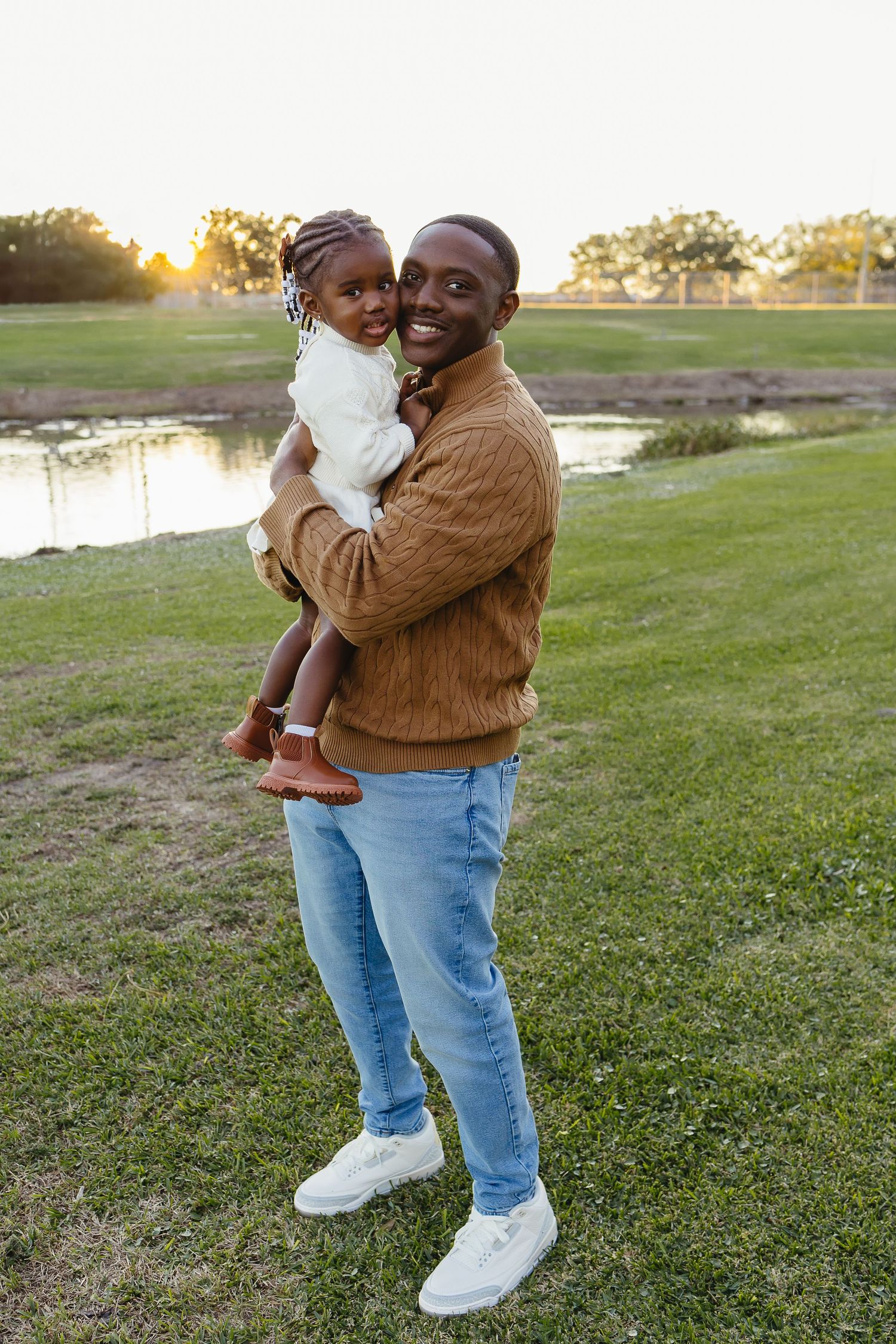 Smiling Black father holding young daughter in white outfit, standing on green grass near pond at golden hour.