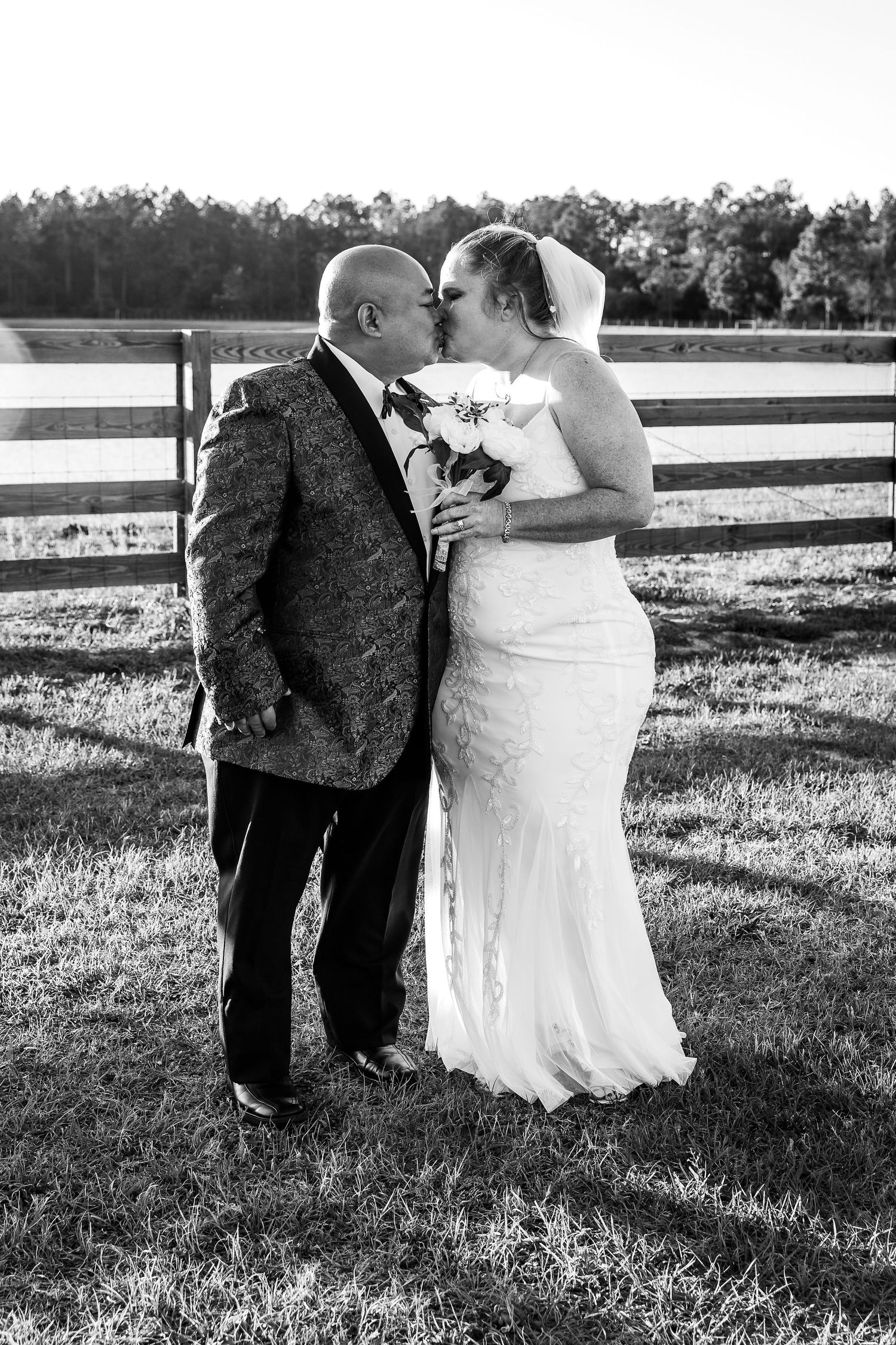 Black and white photo of bride and groom sharing a kiss in a sunlit field with white fence backdrop.