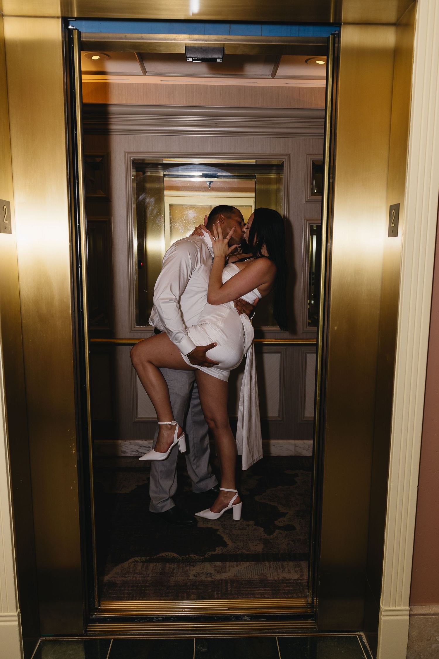 Couple sharing passionate kiss in gold elevator, woman lifted slightly, both dressed in white and gray attire.