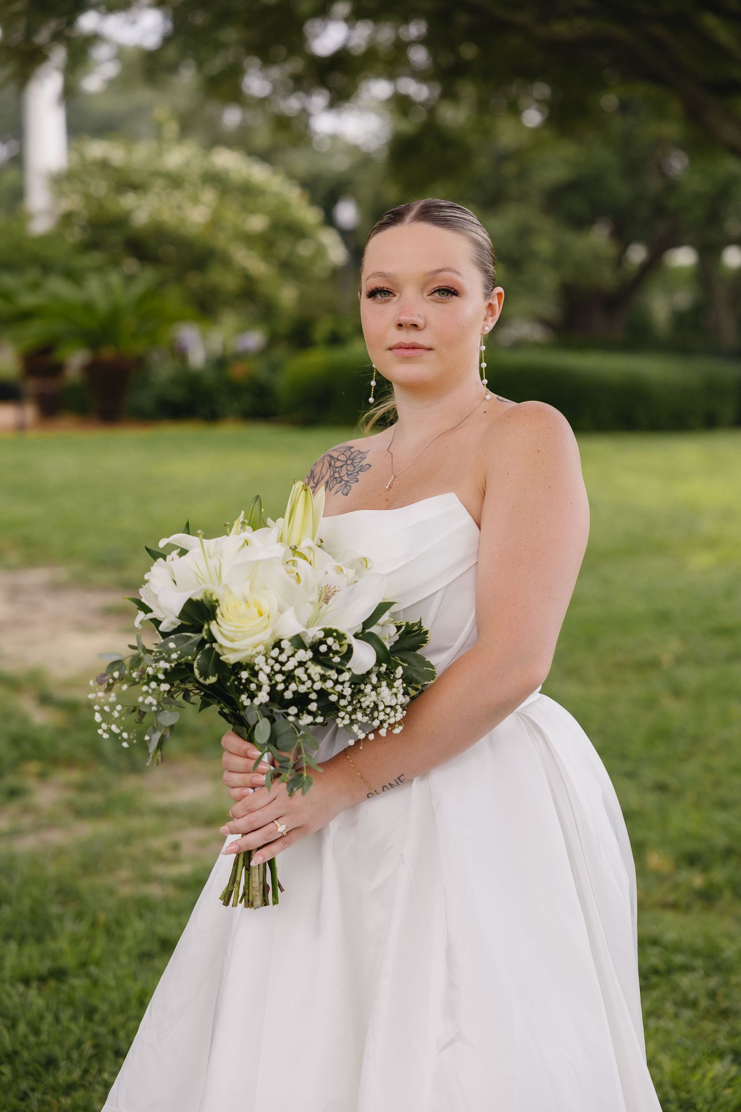 Beautiful bride in strapless white gown holding white lily and rose bouquet in a lush garden setting.