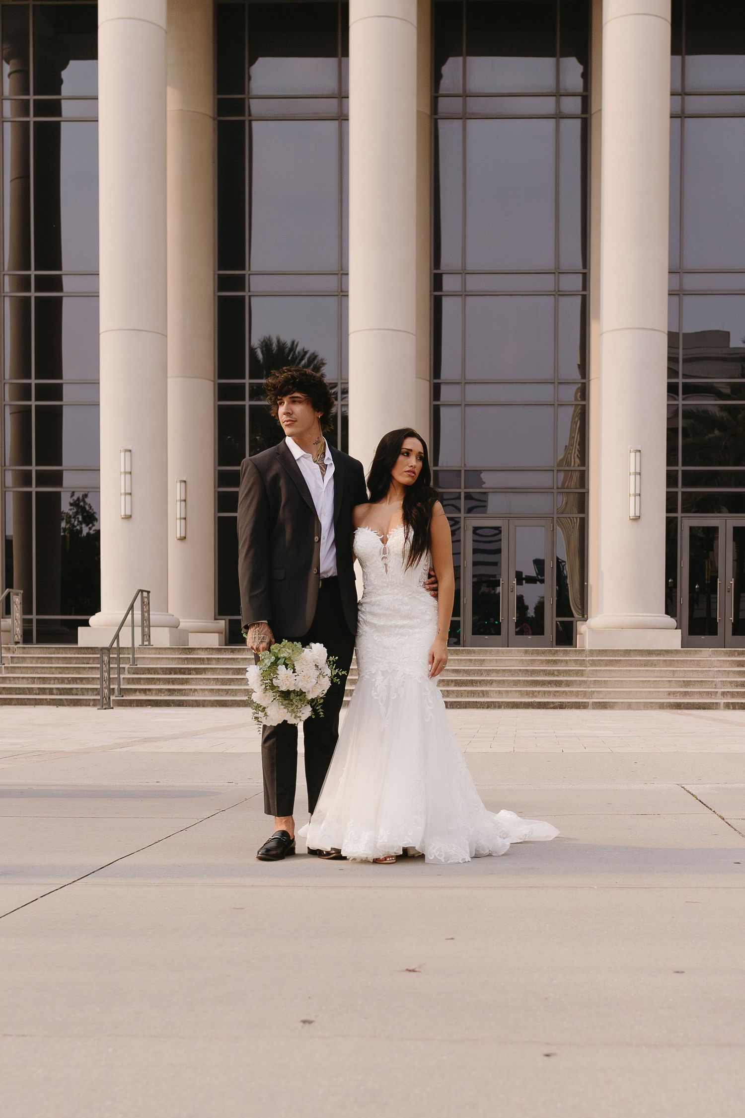 Bride and groom stand before grand columned building, bride holding white floral bouquet.