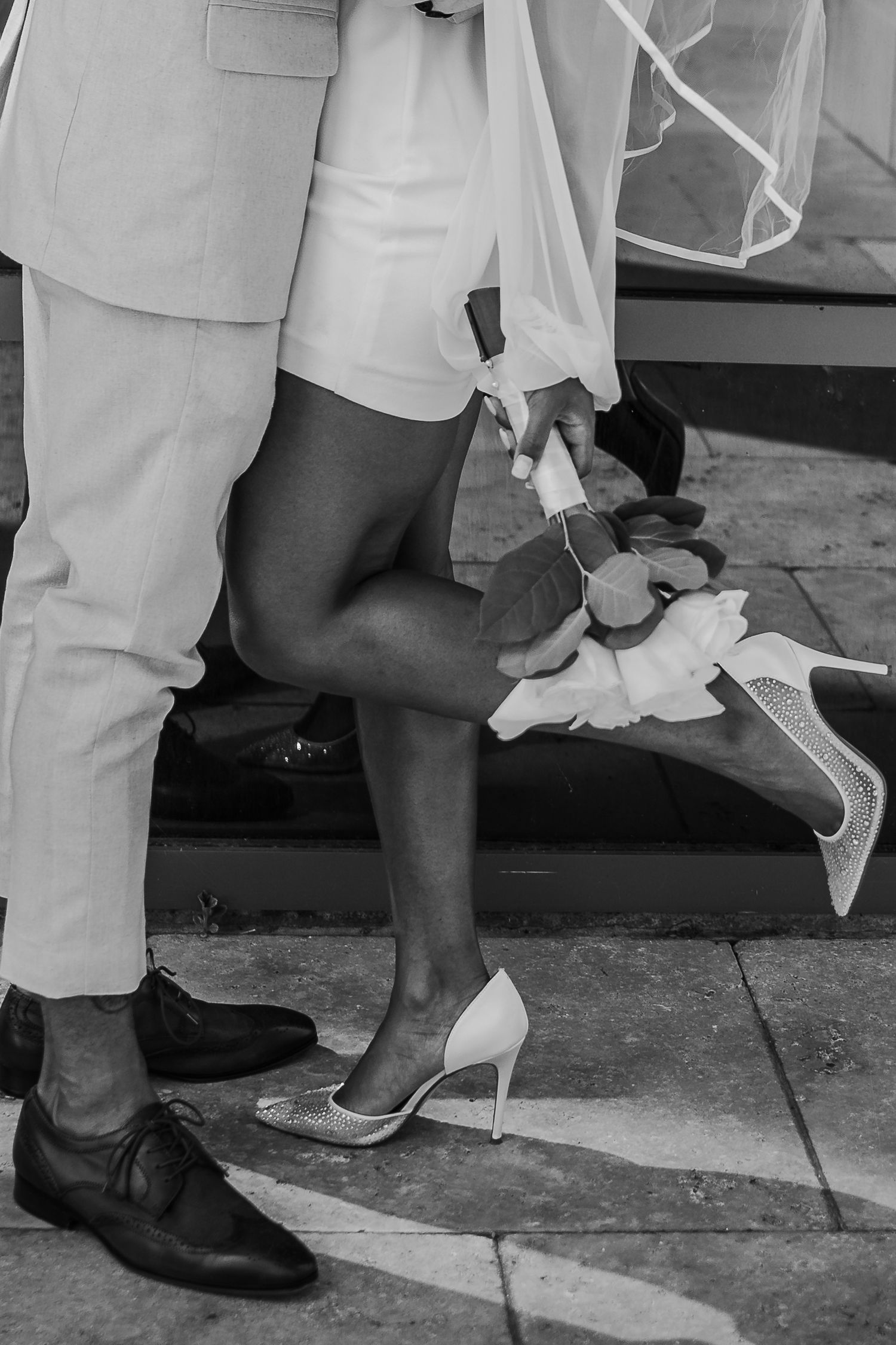 Black and white close-up of bride adjusting her white heels while groom stands beside her in dress shoes.