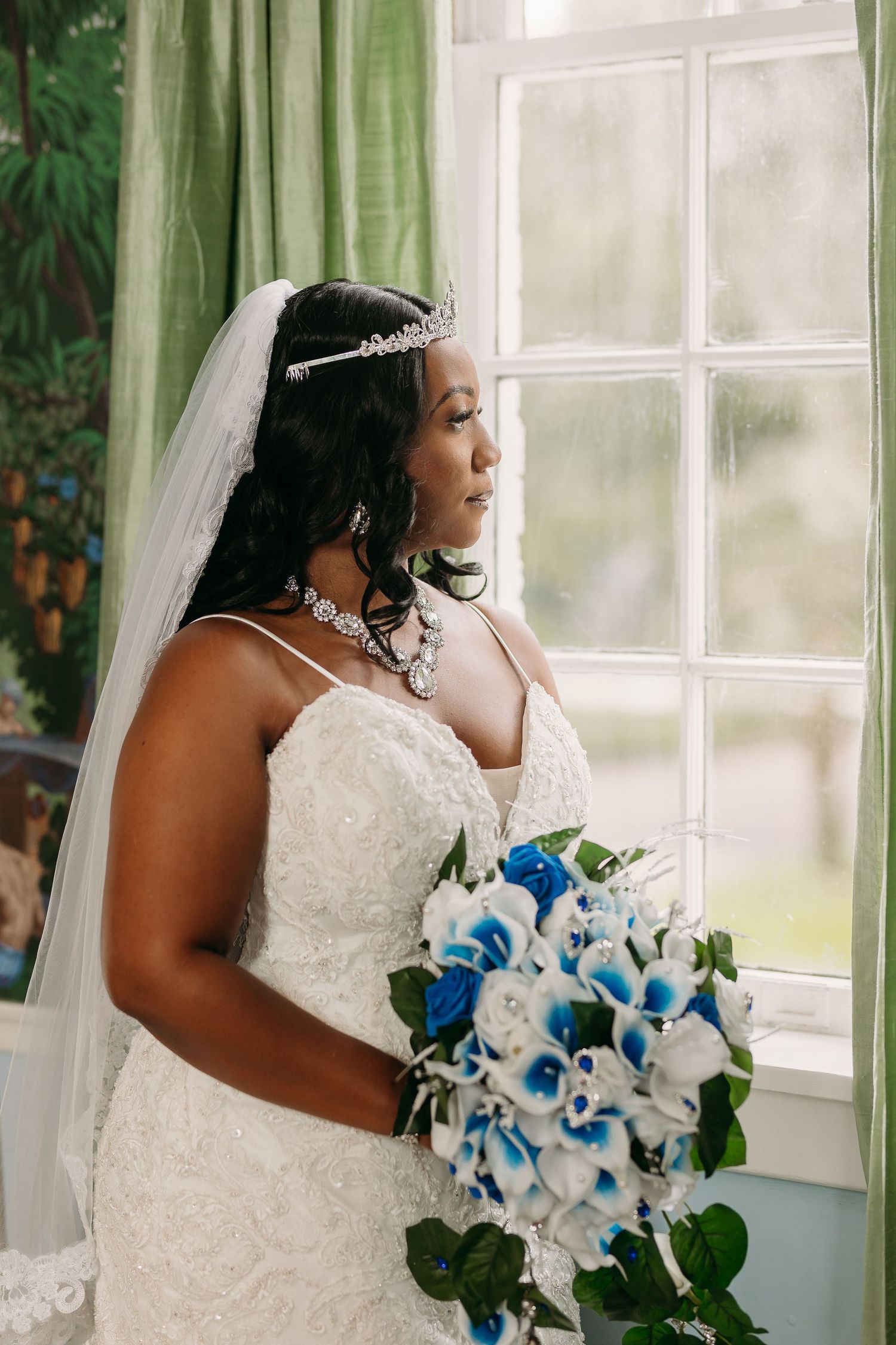 Beautiful Black bride in lace gown holding blue and white bouquet by window.