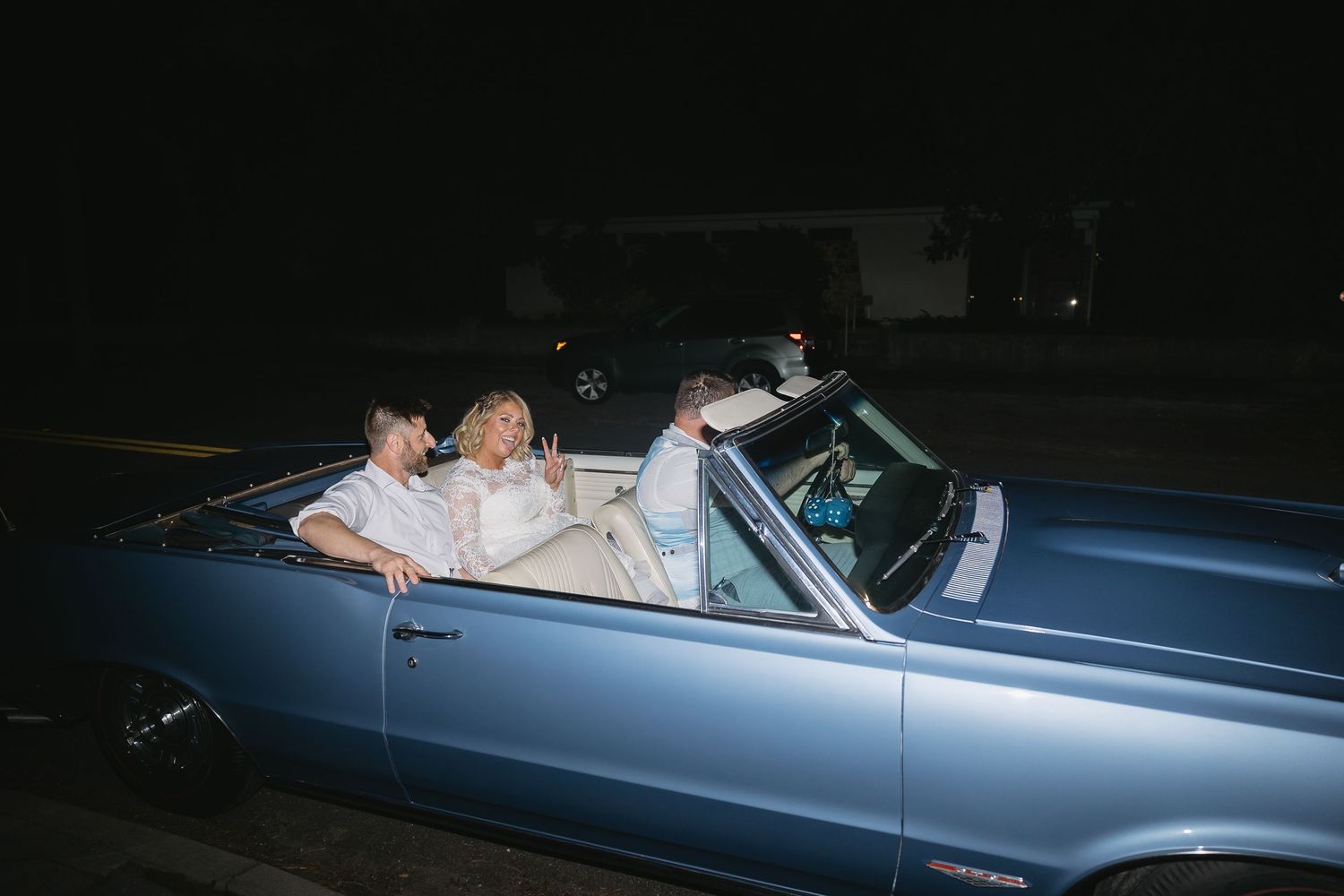Bride and groom riding in vintage blue convertible car at night after wedding.
