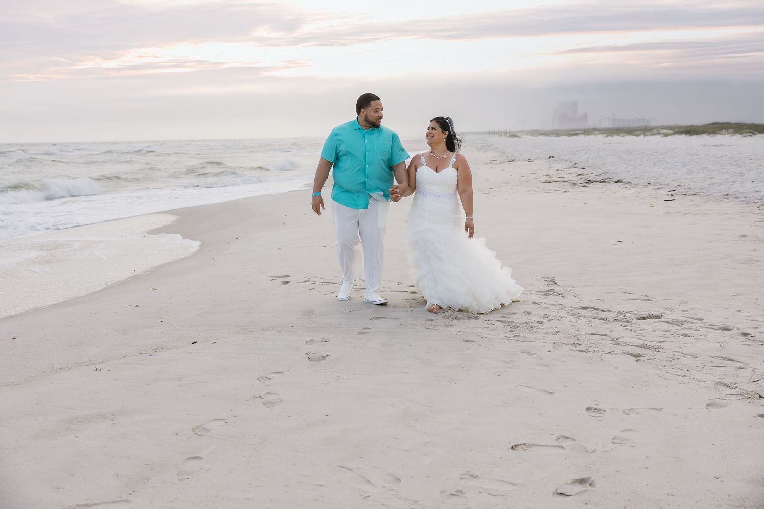 Newlywed couple in wedding attire holding hands walking along ocean shoreline at sunset.