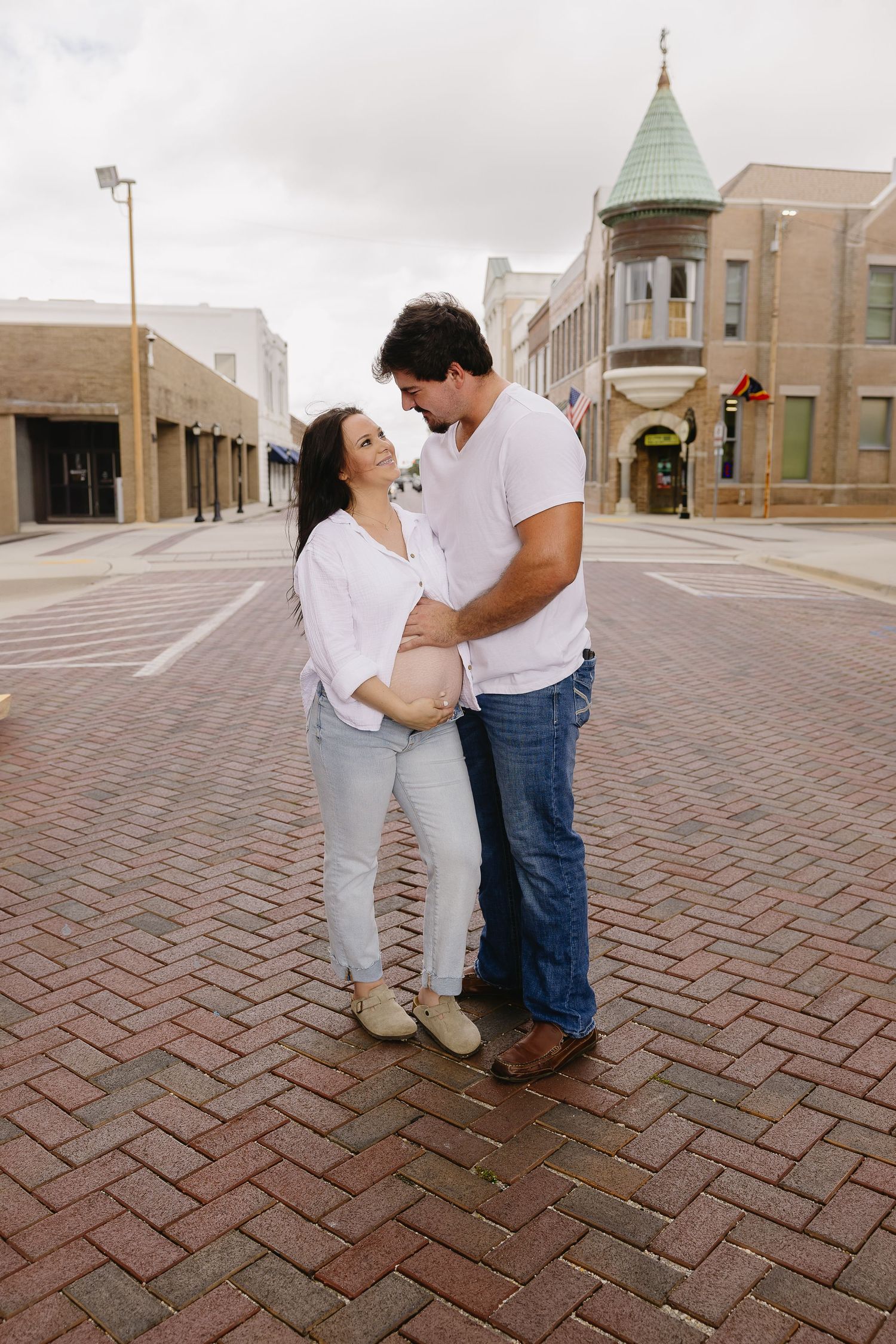 Smiling pregnant couple embracing on a brick plaza, woman in white shirt and jeans, man in blue jeans.