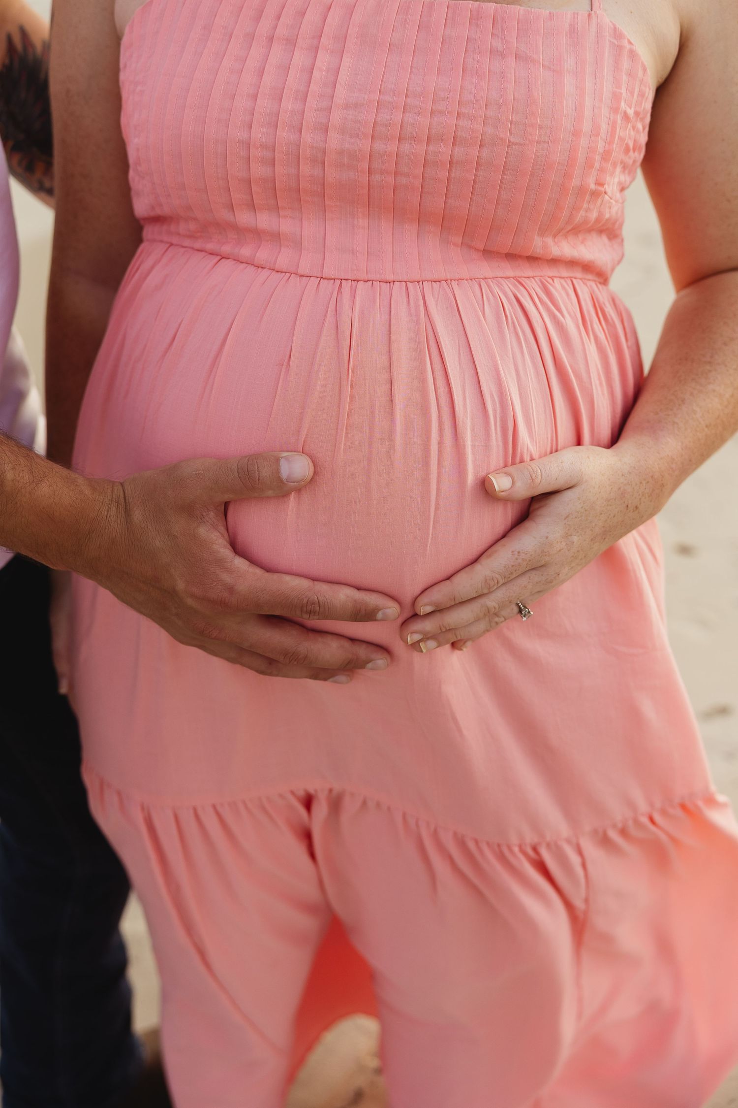 Close-up of couple cradling pregnant belly in pink maternity dress, hands forming heart shape.