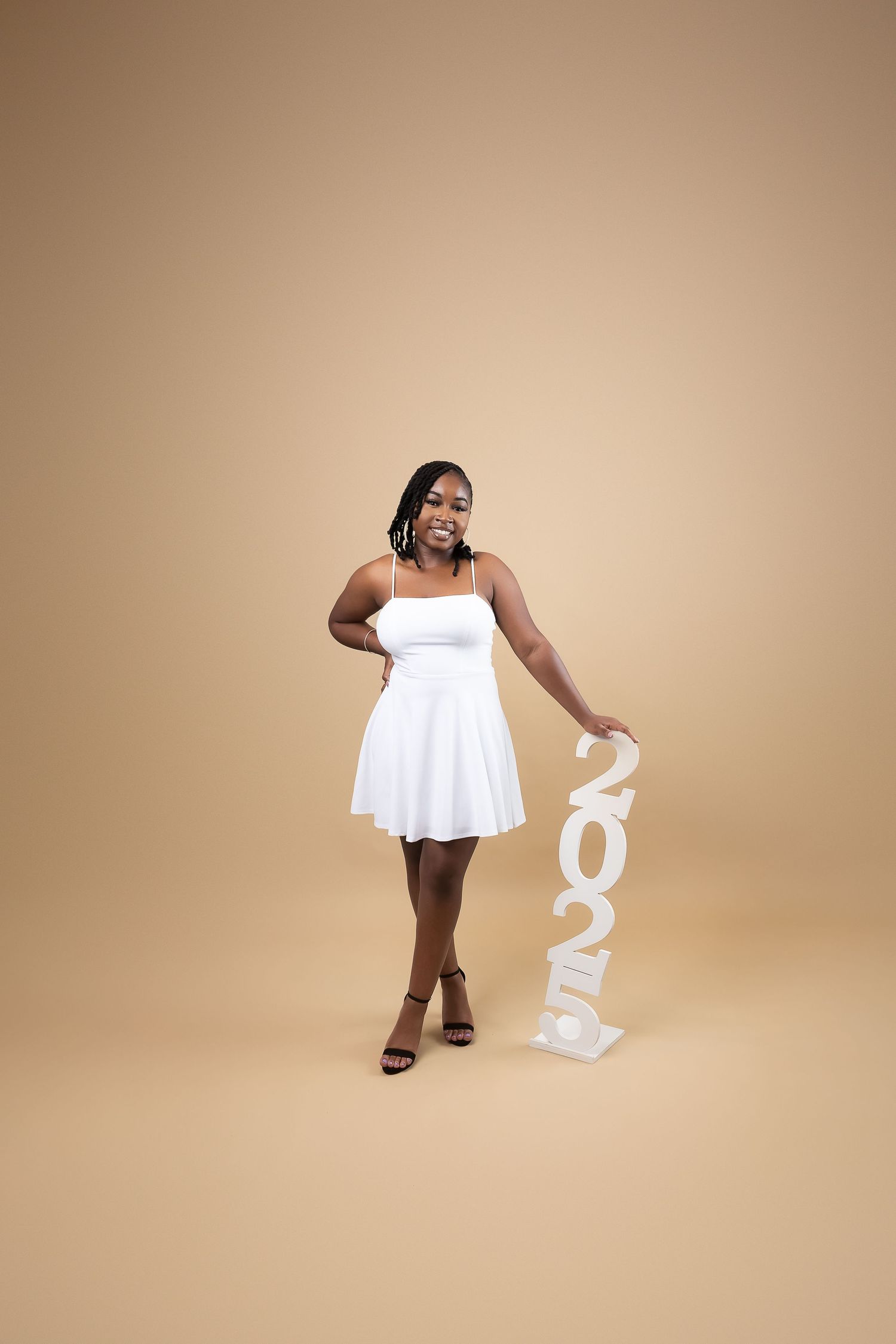 A young Black woman in a white mini dress poses with a 2025 sign on a beige background.