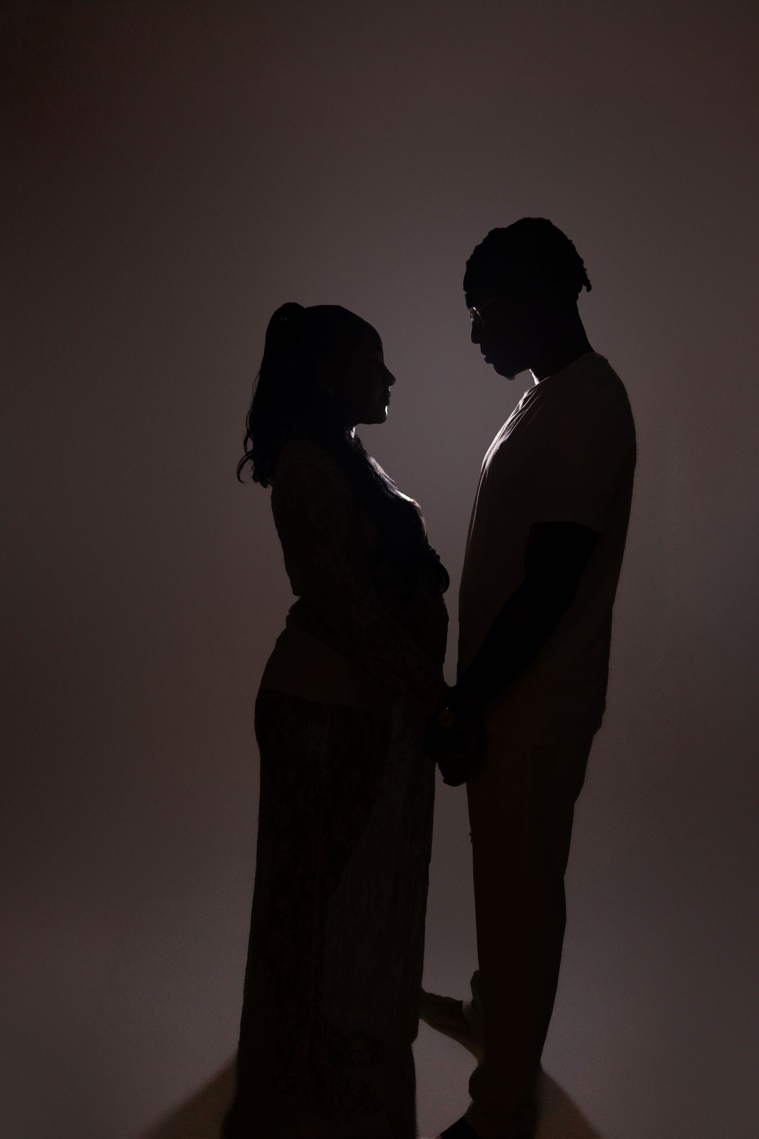 Romantic silhouette of pregnant couple holding hands facing each other against dark background.