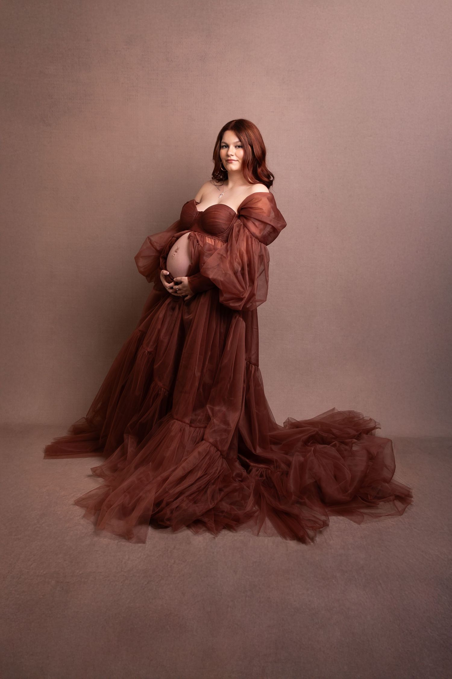 Pregnant woman in elegant rust-colored off-shoulder tulle gown poses in studio on brown background.