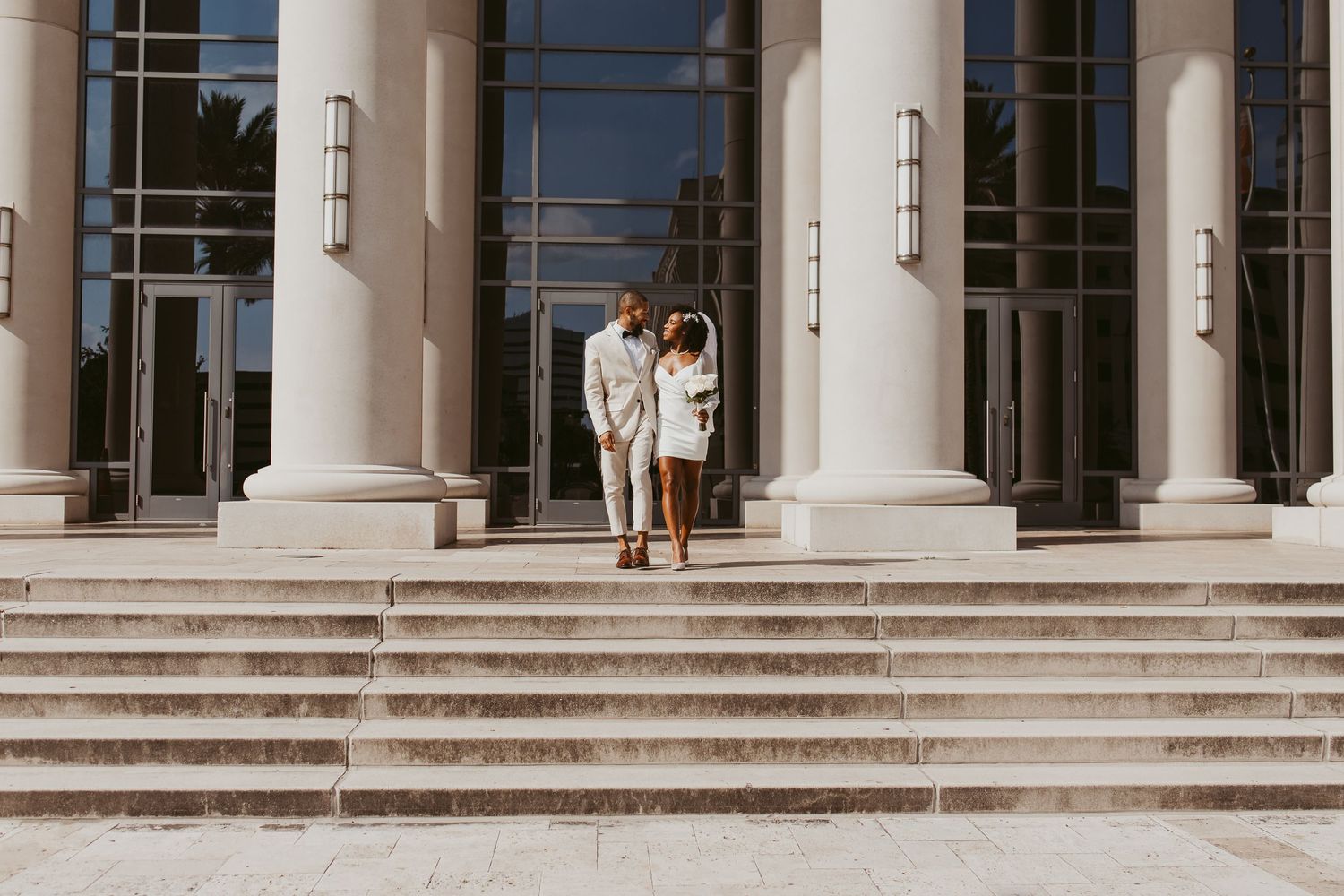 Bride and groom walking hand in hand down steps of a grand building with tall white columns.