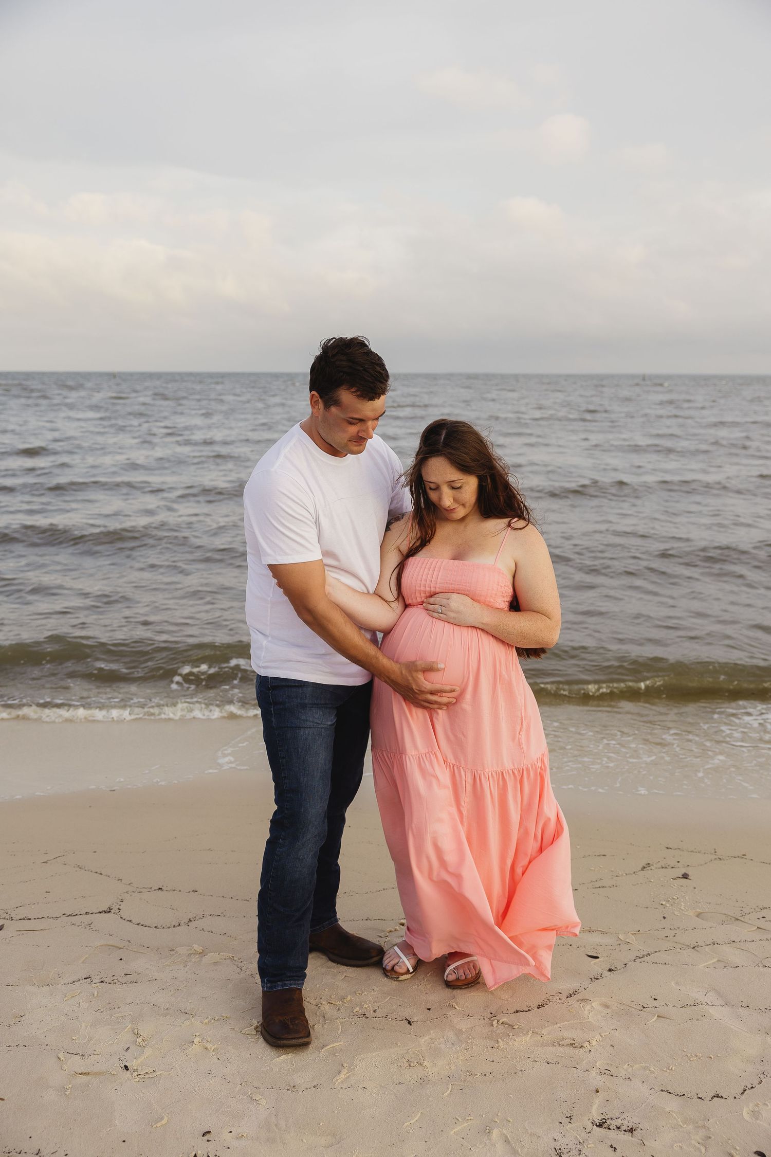 Couple cradling pregnant belly on beach, man in white tee and jeans, woman in pink maxi dress, ocean backdrop.