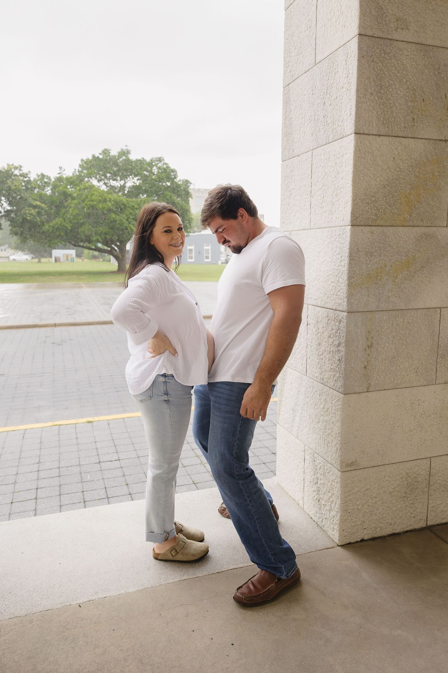 Pregnant woman and partner posing by a stone column, both in white tops and jeans outdoors.