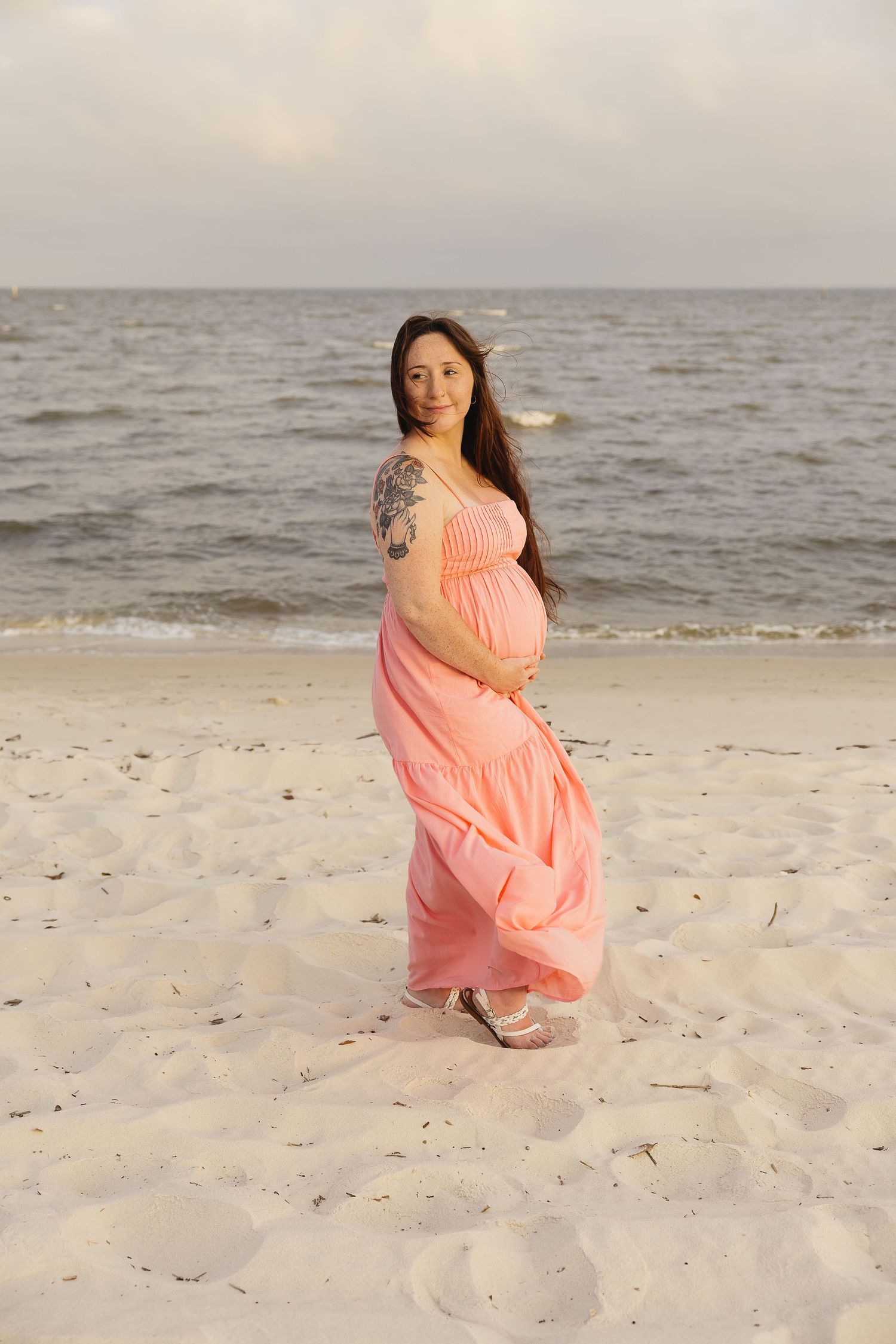 Smiling pregnant woman with tattoo in pink beach dress standing on sandy shore by ocean.