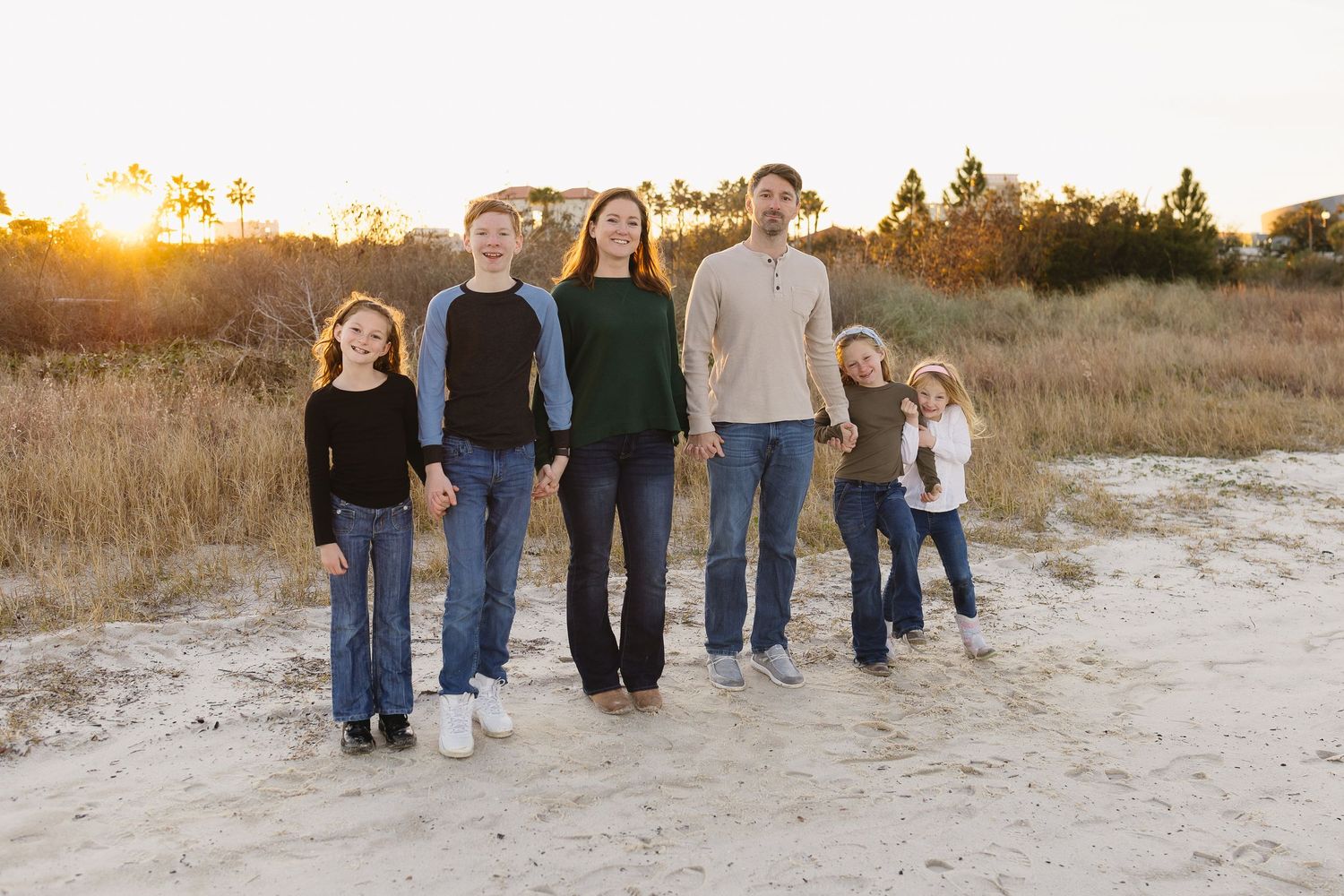 A family of six stands together on a sandy beach path surrounded by golden grass at sunset.