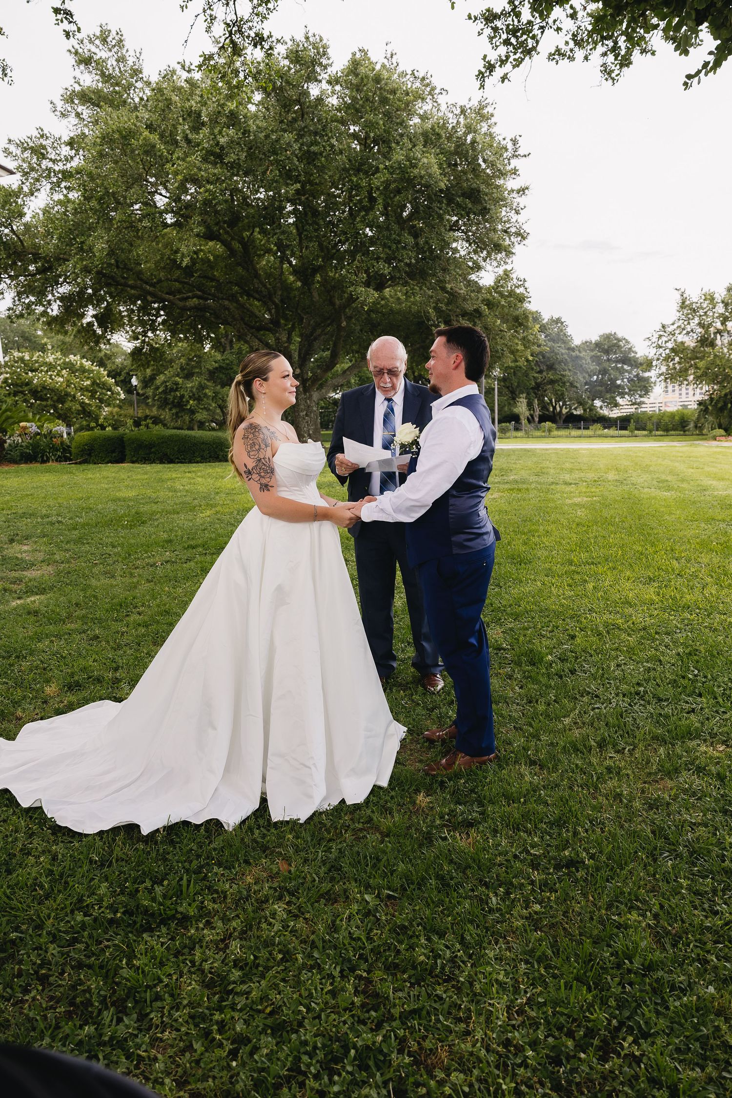 Bride and groom exchanging vows with officiant on green lawn under large oak tree.