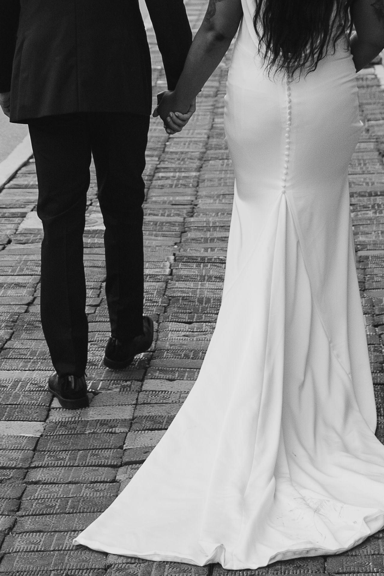 Black and white photo of bride and groom holding hands walking on brick path.