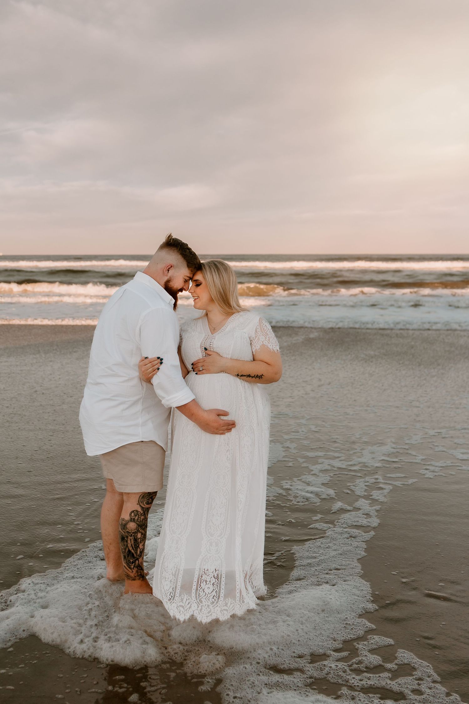 Pregnant couple embracing at sunset on beach, man in white shirt, woman in white lace maternity gown.