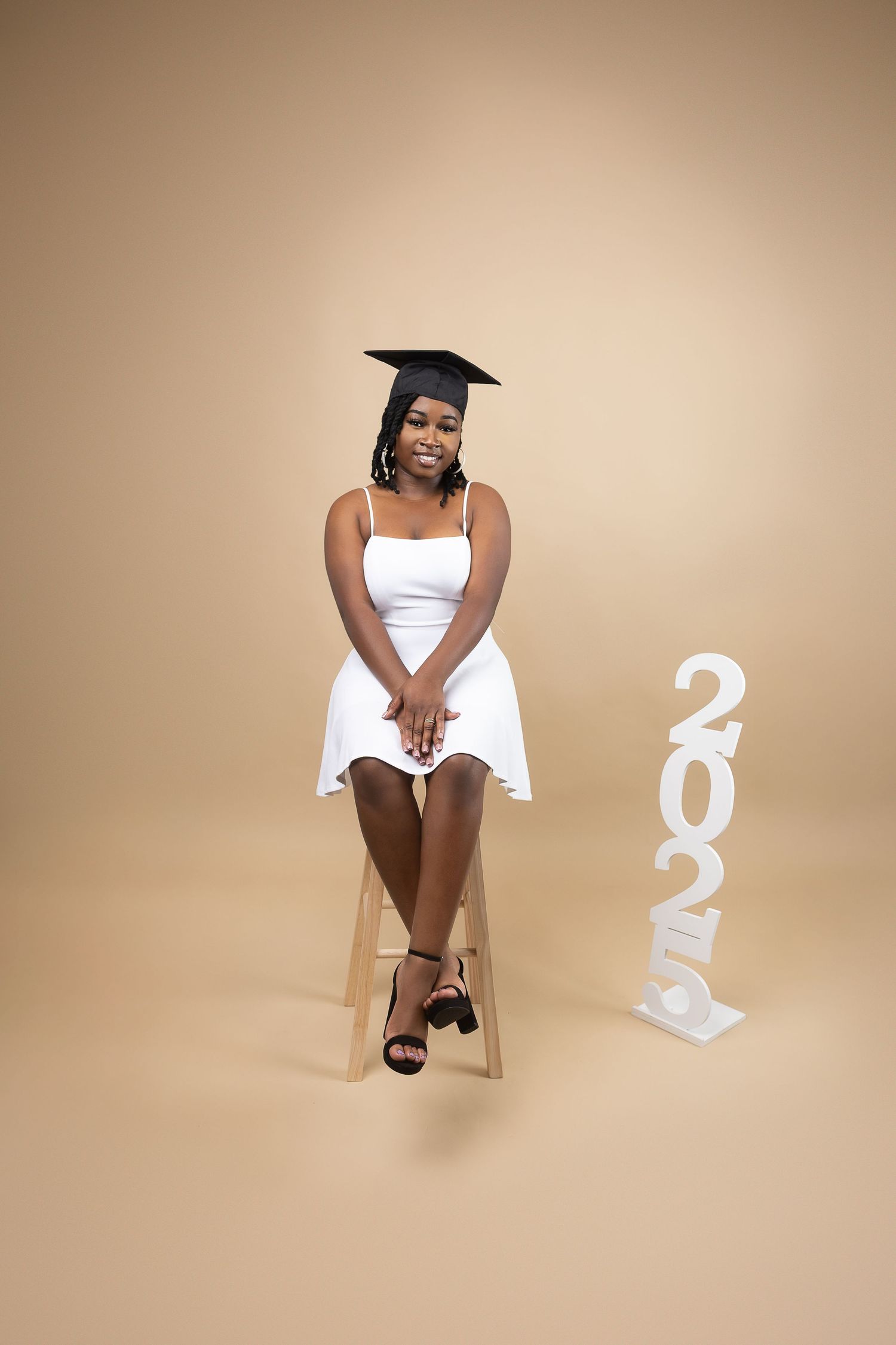 Young woman in white dress and graduation cap seated beside 2025 sign on beige backdrop.