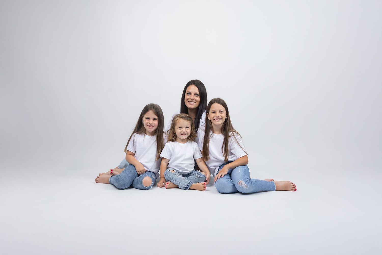 Mother and three daughters in white shirts and jeans sit together smiling on white studio background.