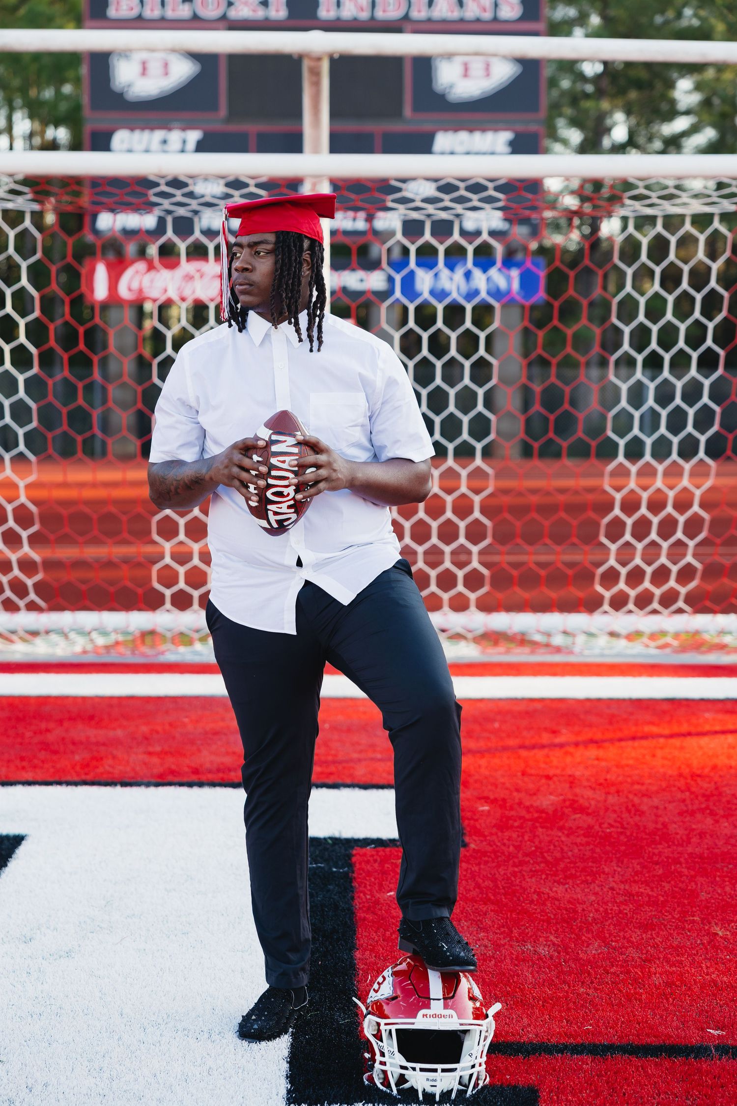 Senior athlete in white shirt and red graduation cap posing with football on Biloxi Indians field.